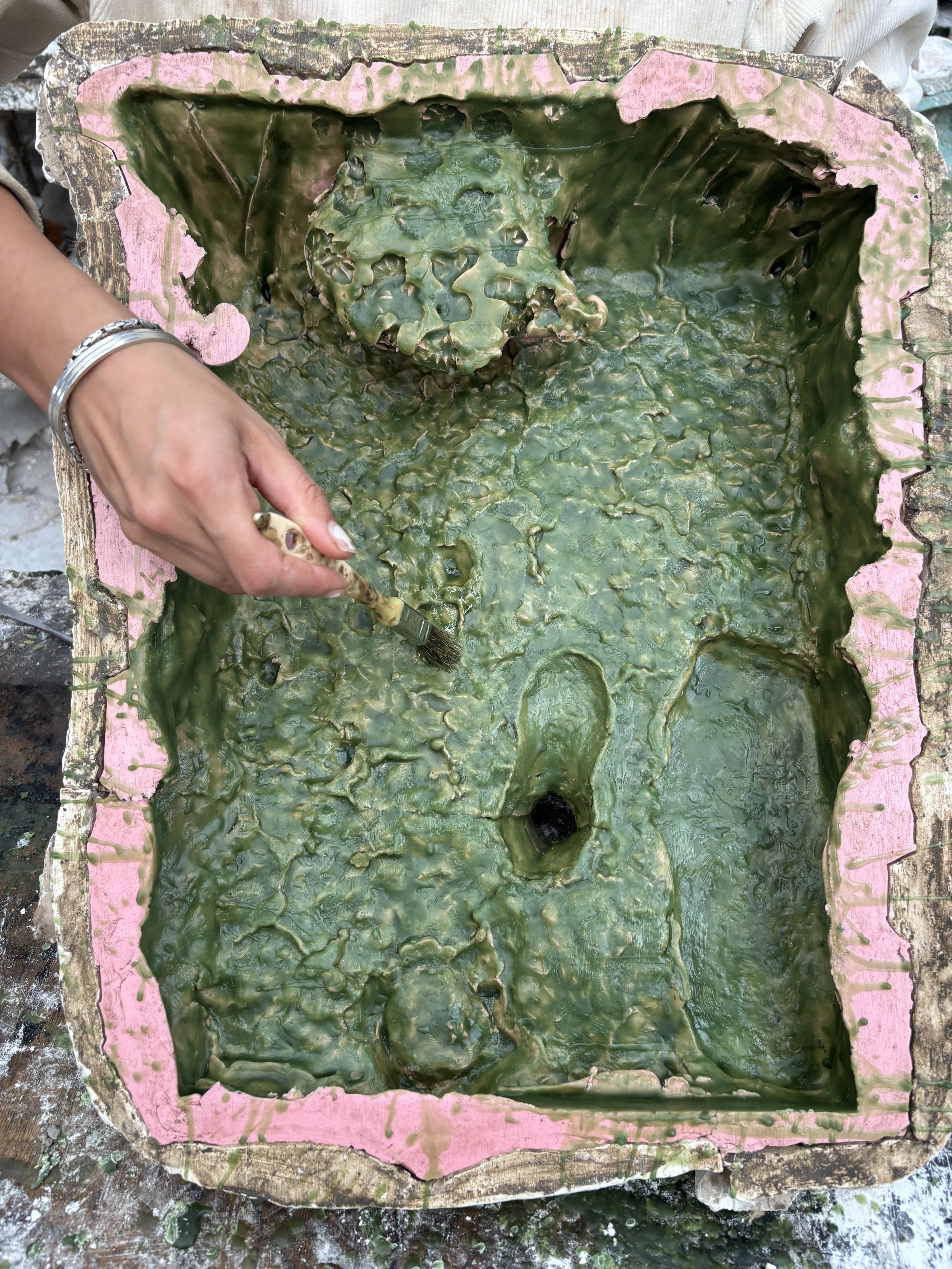 A person holding a paintbrush working on the green wax coating inside a mold for making sculpture, with pink silicone around the mold edges.
