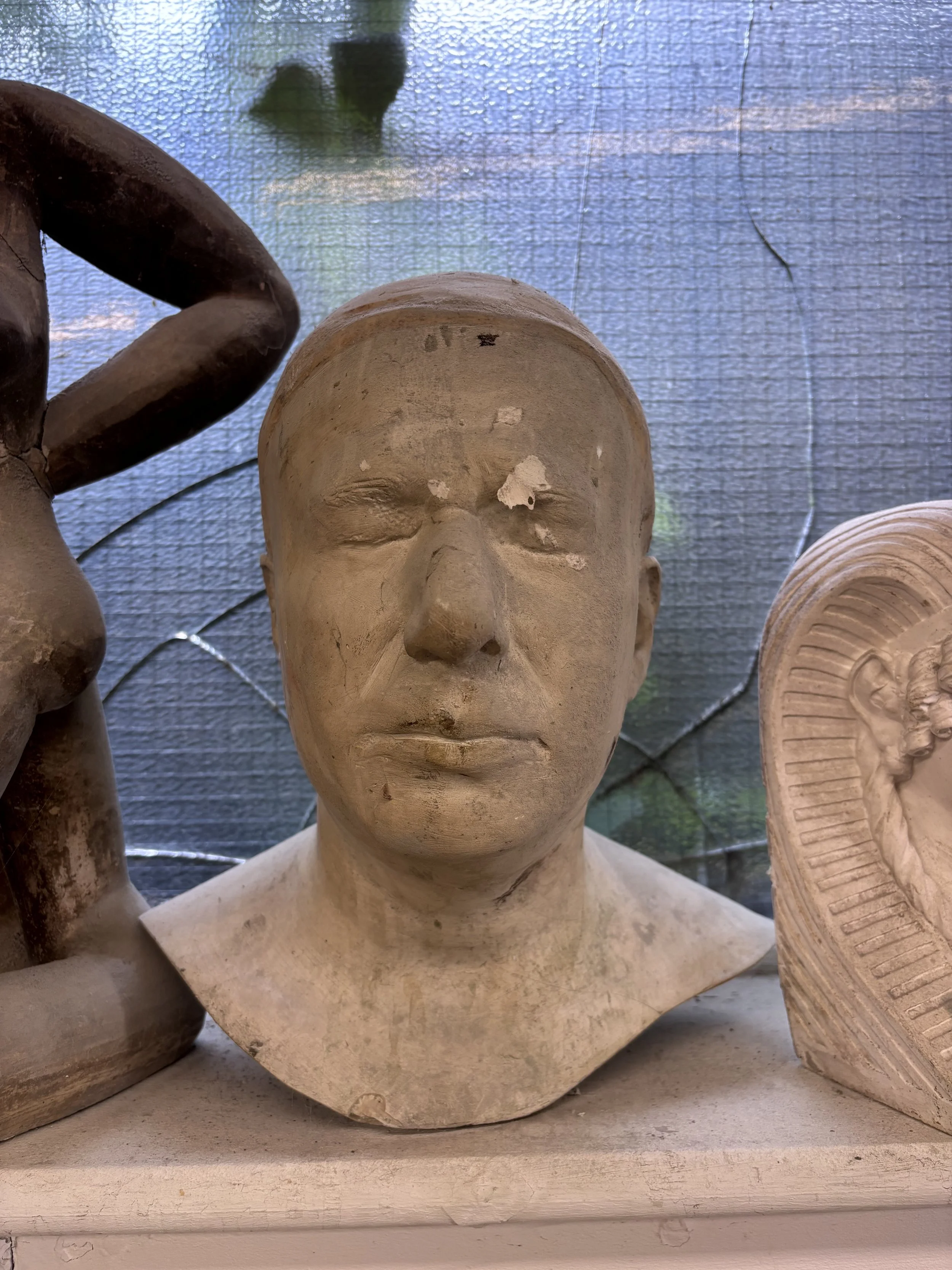 A sculpture of a human head with closed eyes, featuring a calm facial expression, placed among other sculptures on a surface.