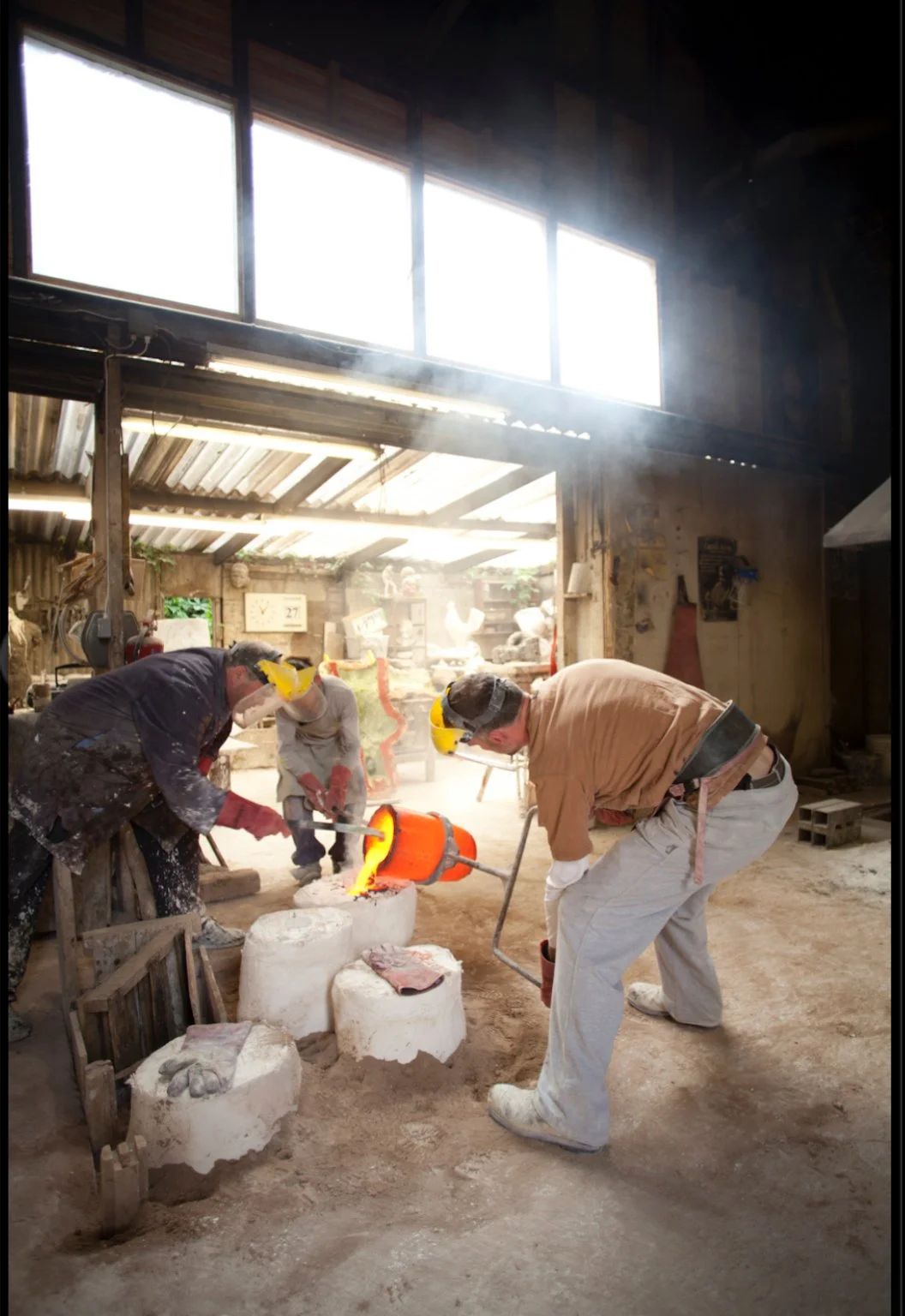 Three artisans working with a crucible and glowing hot metal inside a rustic workshop foundry, illuminated by sunlight streaming through large windows.