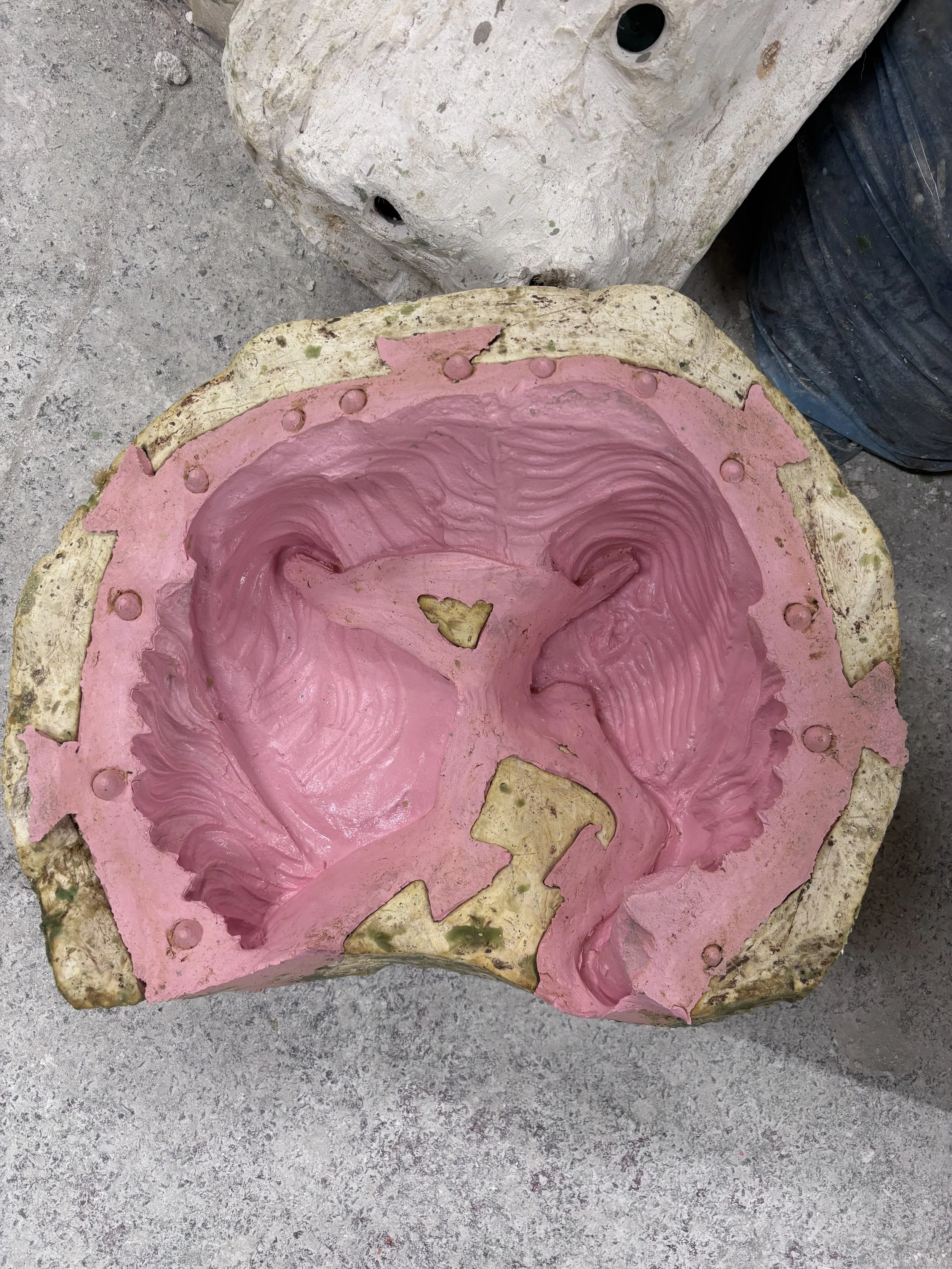 A pink rubber mold with a heart shape and swirling details inside, resting on a rough concrete surface, with a large white stone and a black container nearby.