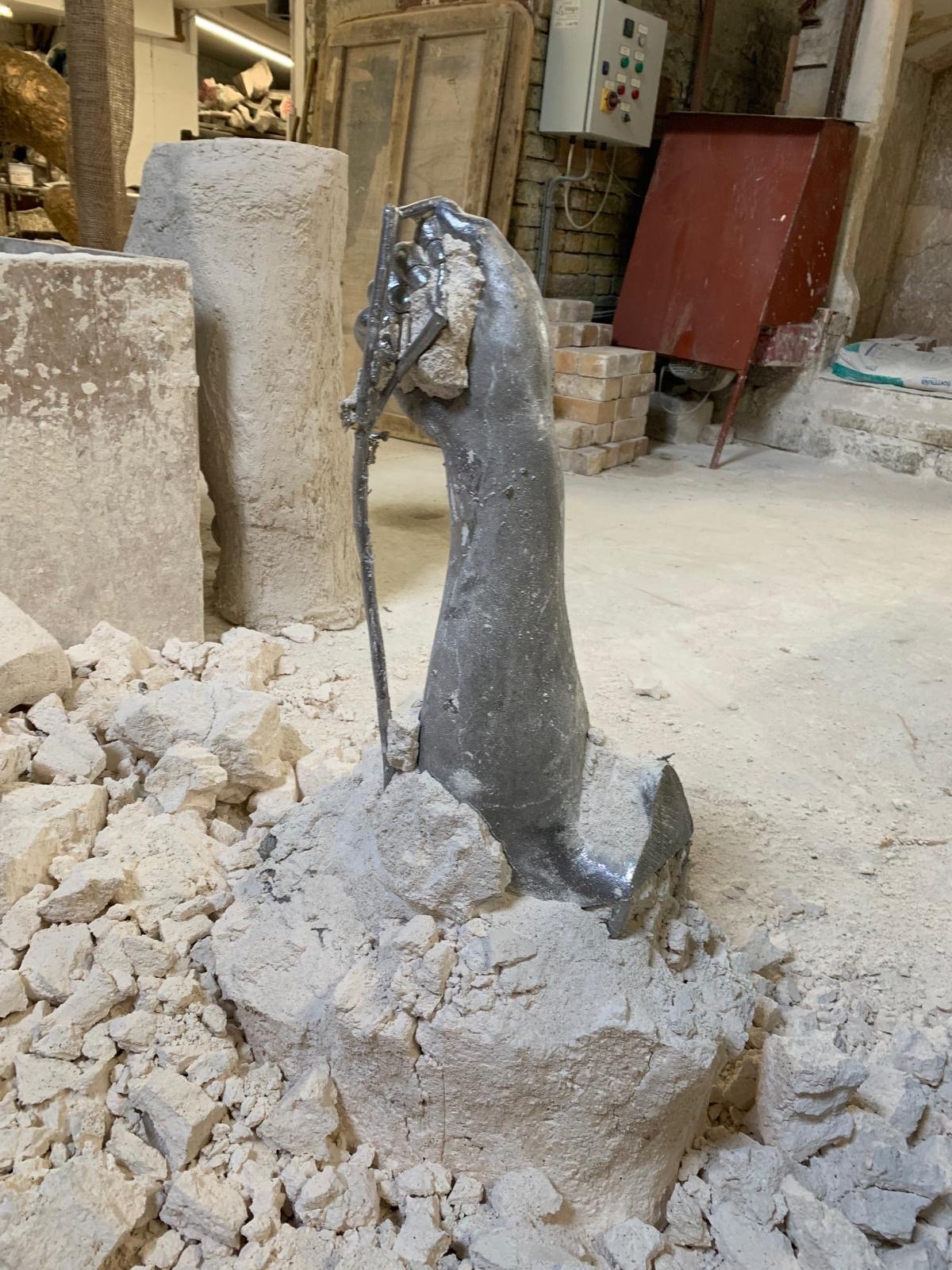 A lead sculpture of an arm and hand, standing among smashed plaster in a foundry with tools, bricks, and equipment in the background.