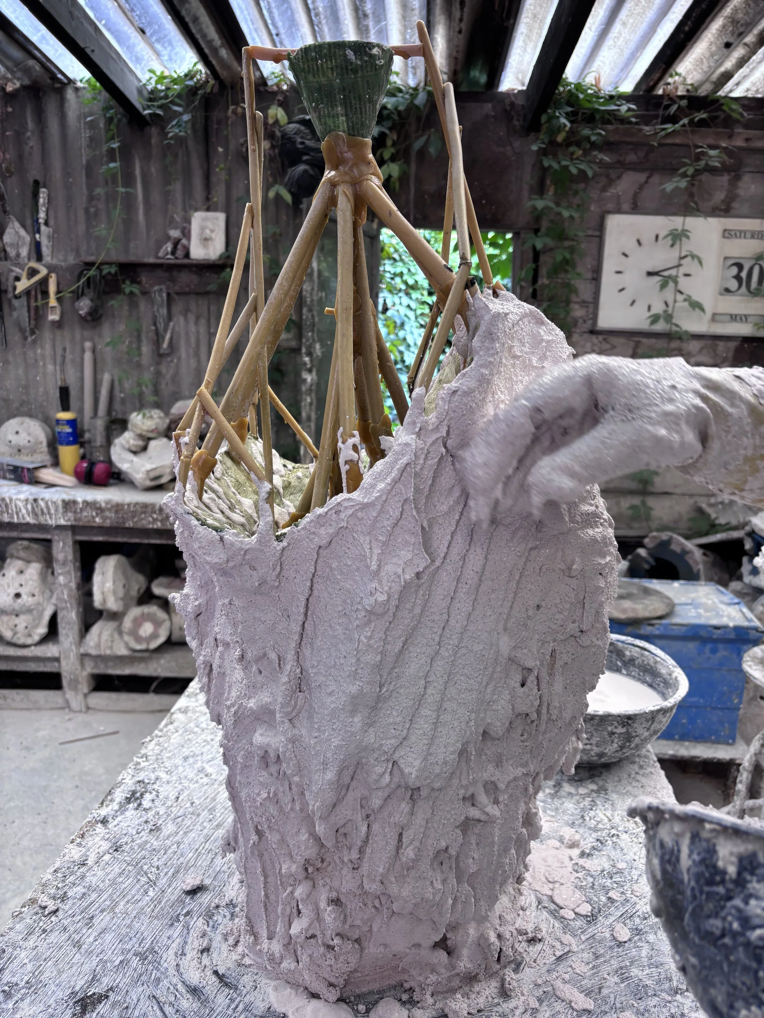 Plaster investment being applied to a wax sculpture with a wax feeding system shown above, part of the lost wax castig process, in a workshop with tools and materials in the background.