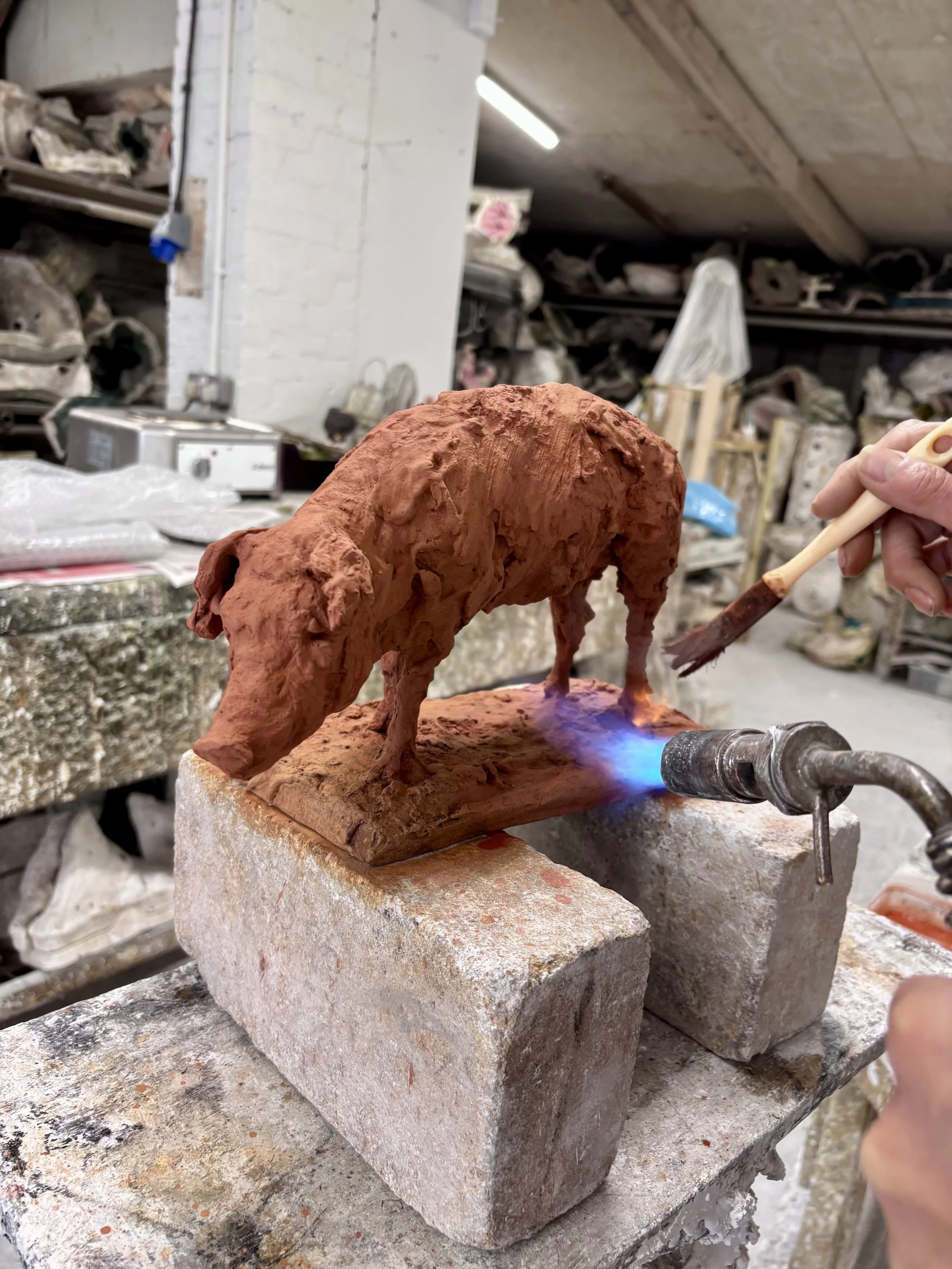 Pig by Nicola Hicks. A person applying patina using a blow torch on a bronze sculpture of a pig using a brush in an artist's workshop.
