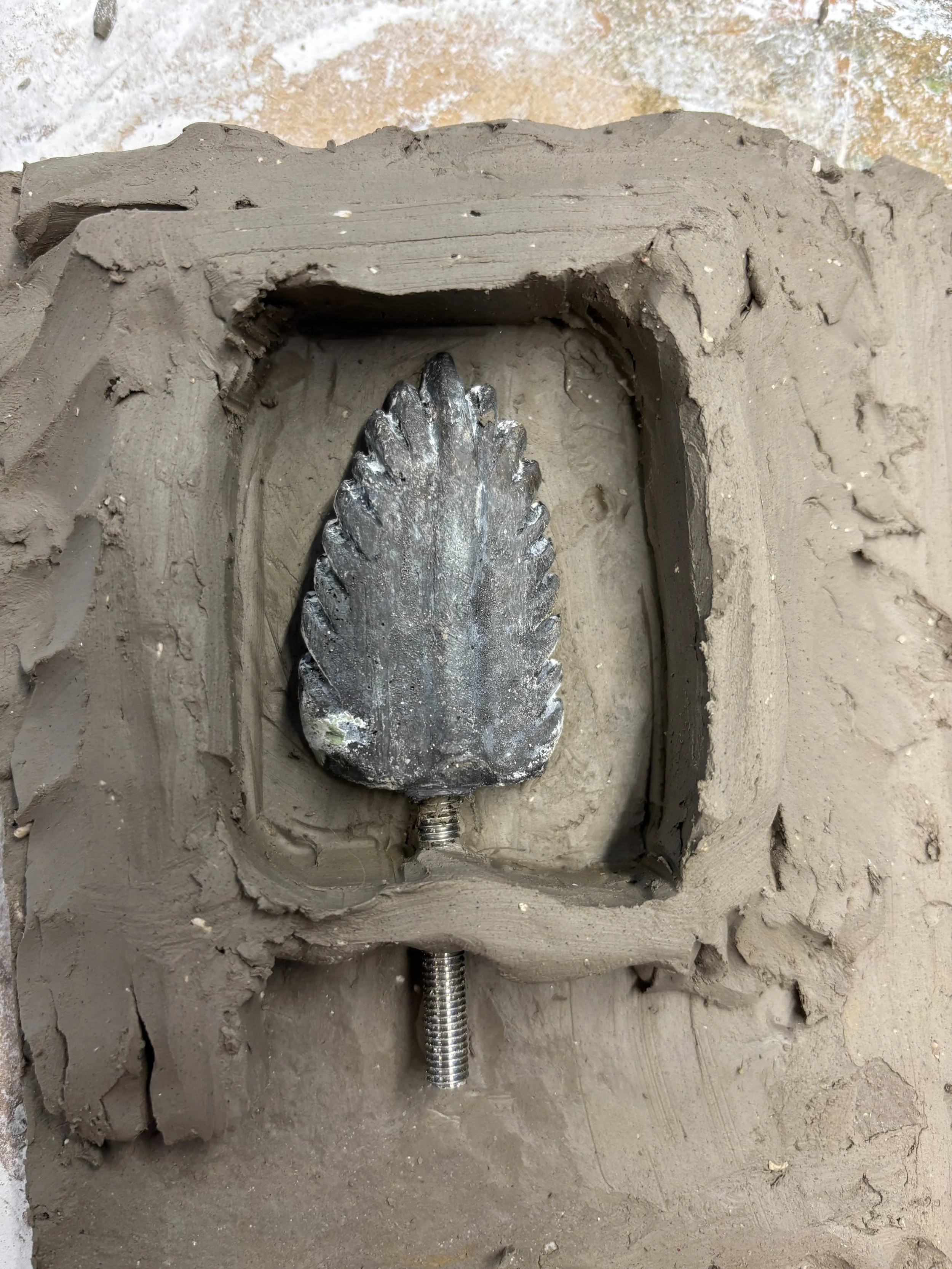 A metal leaf-shaped ornament embedded in a clay wall with a threaded metal rod extending downward.