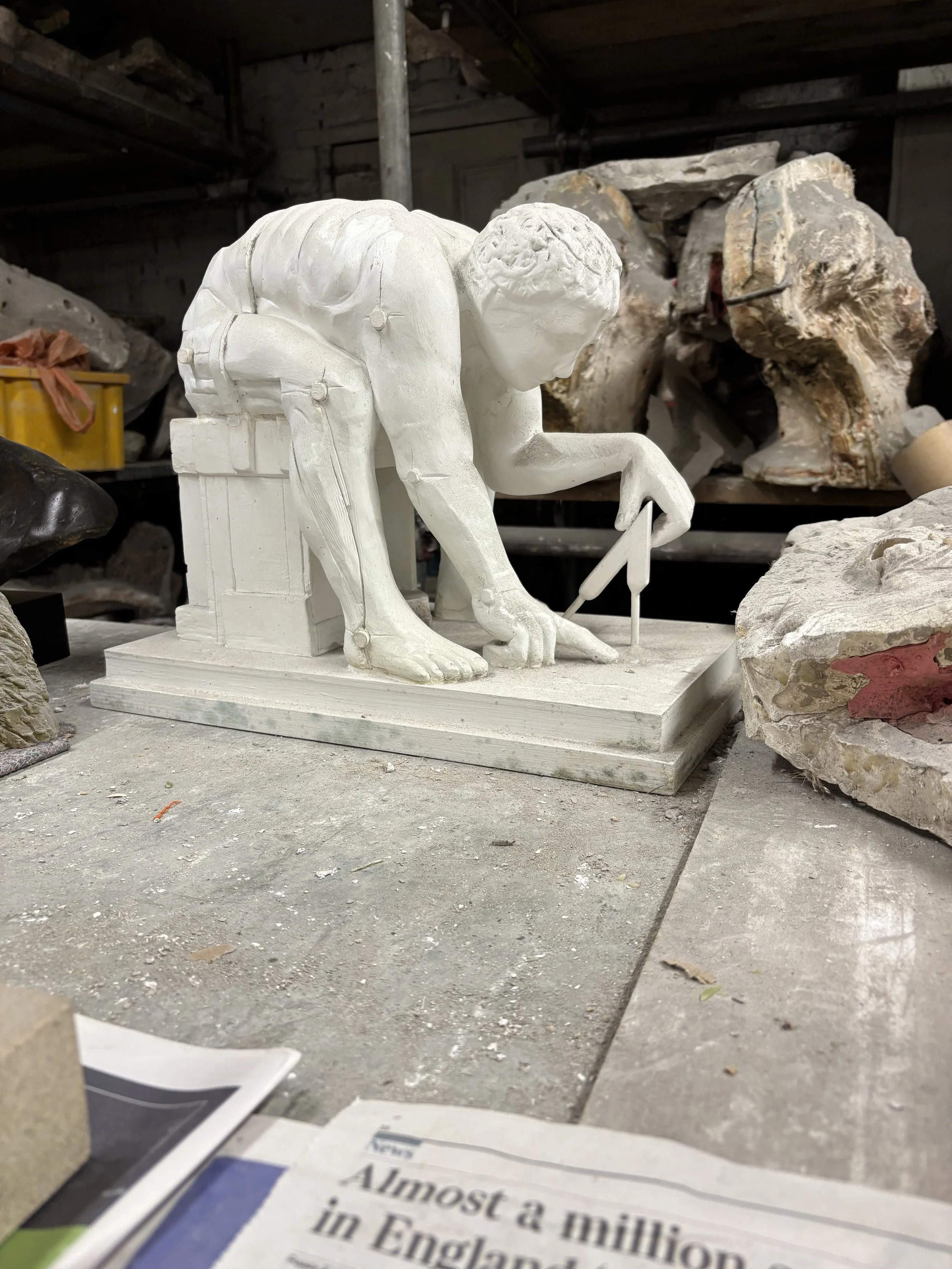 Newton by Eduardo Paolozzi. White plaster sculpture of a man in a crouching position, holding a tool in one hand, placed on a base, surrounded by large rocks or stones in a workshop or art studio.