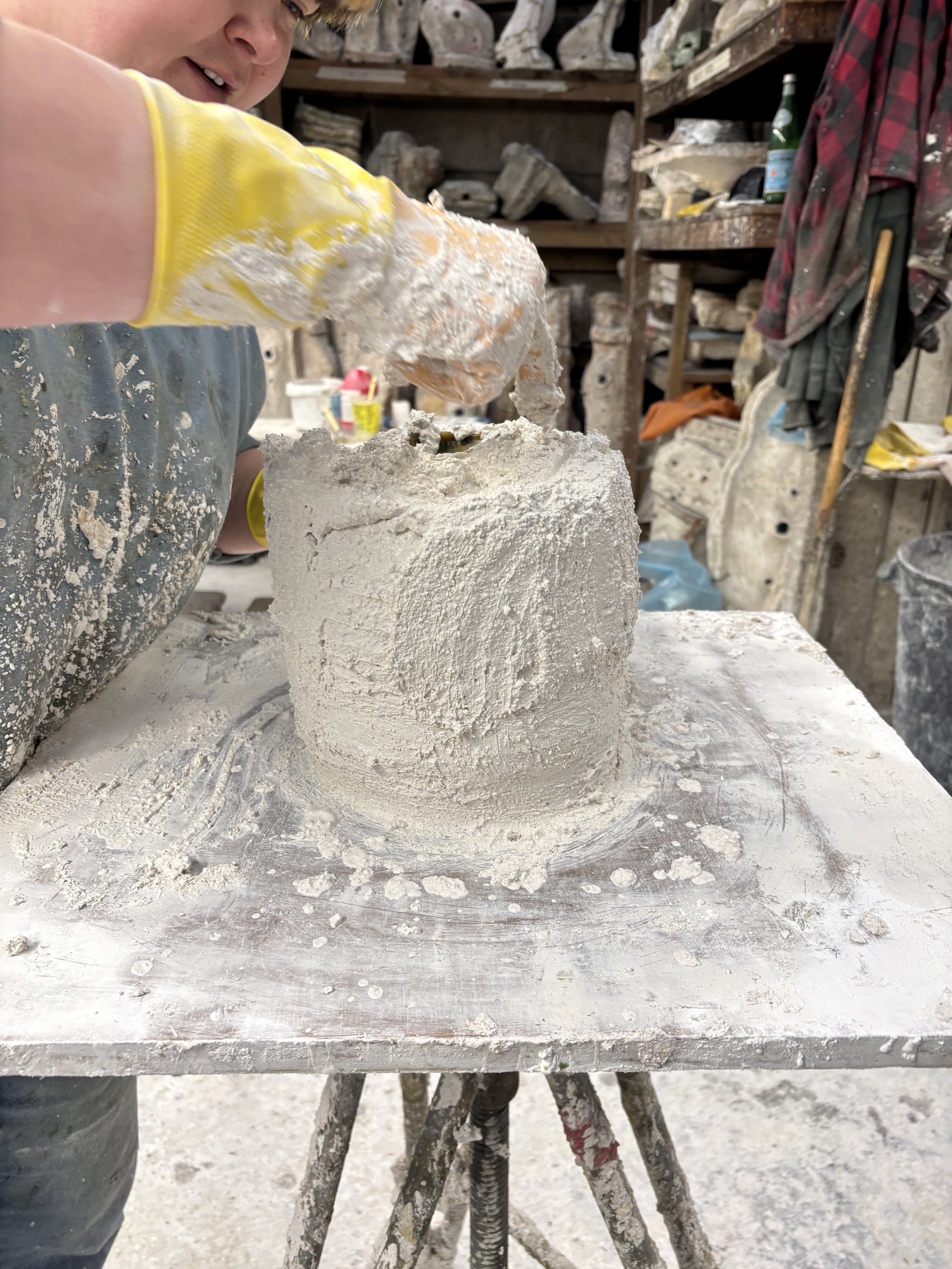 A person applying plaster investment to a wax sculpture in a dusty workspace, with tools, plaster, and sculptures in the background.