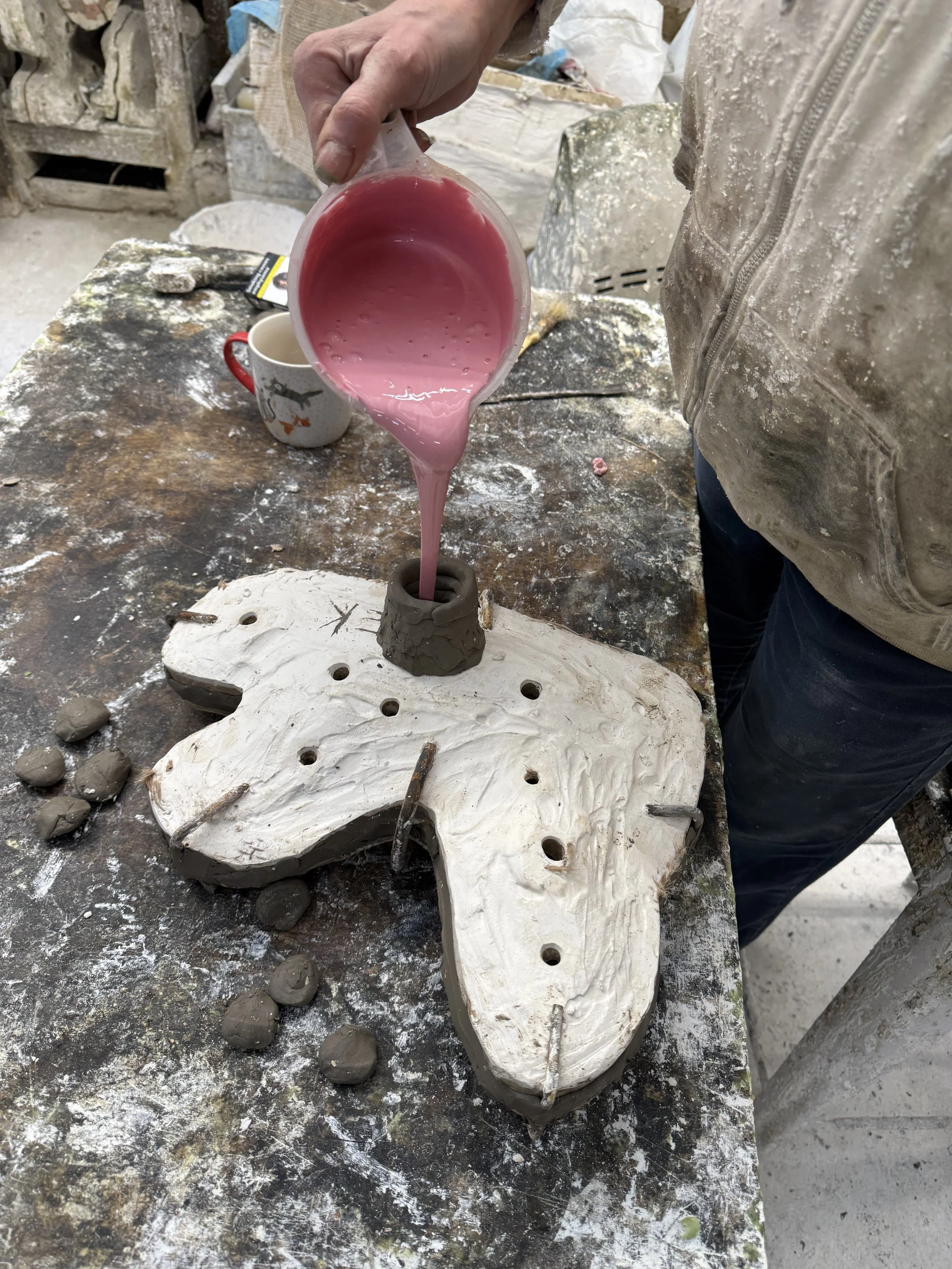 A person pouring pink liquid silicone rubber into a plaster mold on a worktable in a foundry studio.