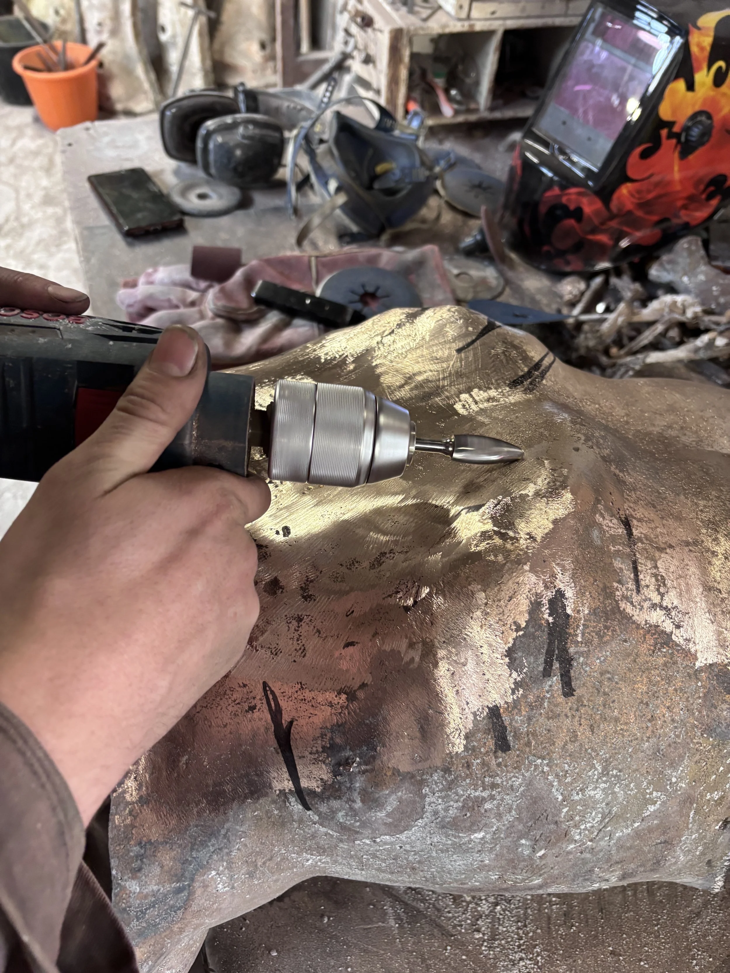 Person holding a rotary tool working on a large piece of metal with a textured, shiny surface, in a workshop with tools and equipment in the background.