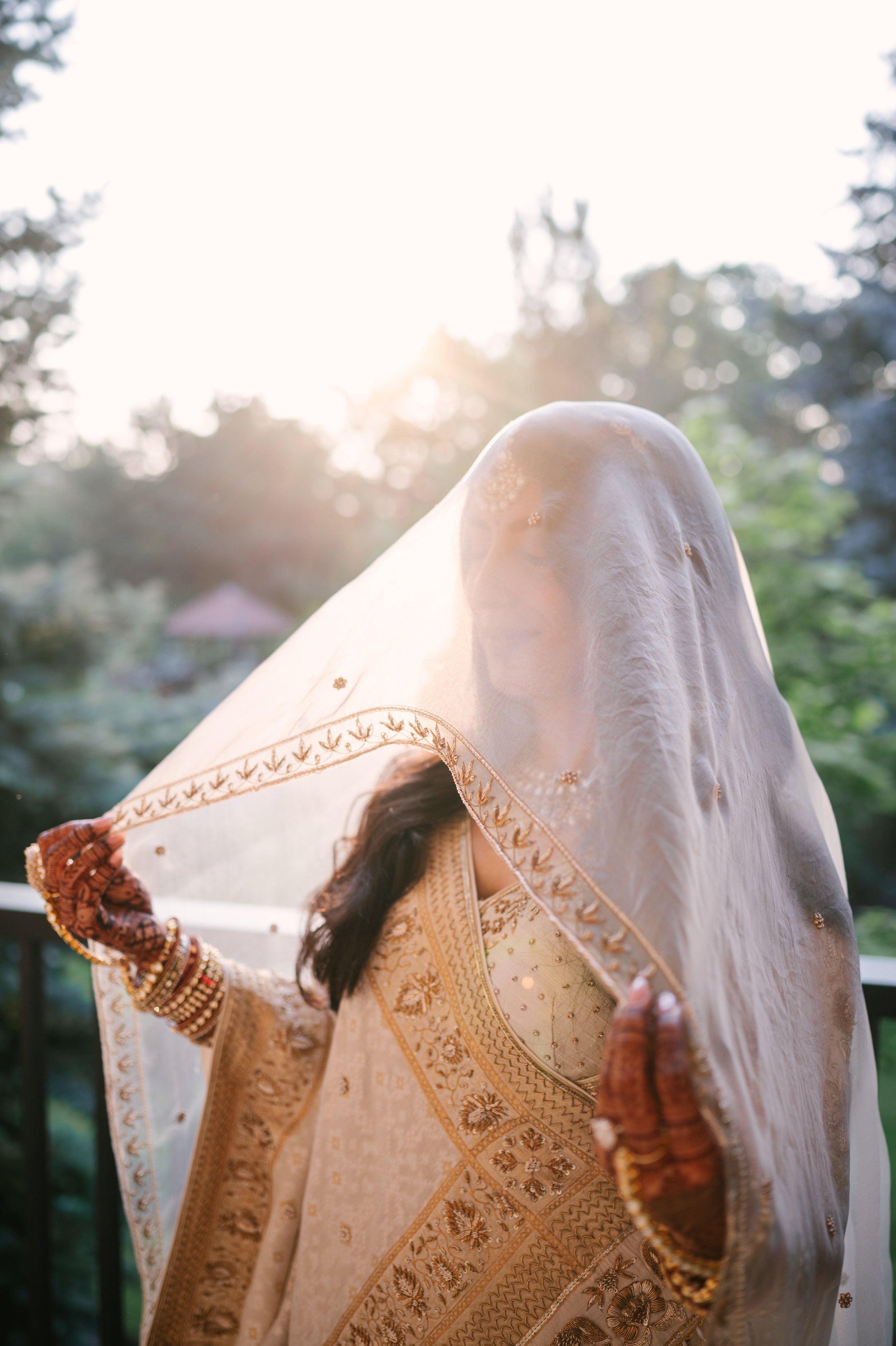 A woman in traditional South Asian attire, draped in a sheer veil, standing outdoors with sunlight in the background.