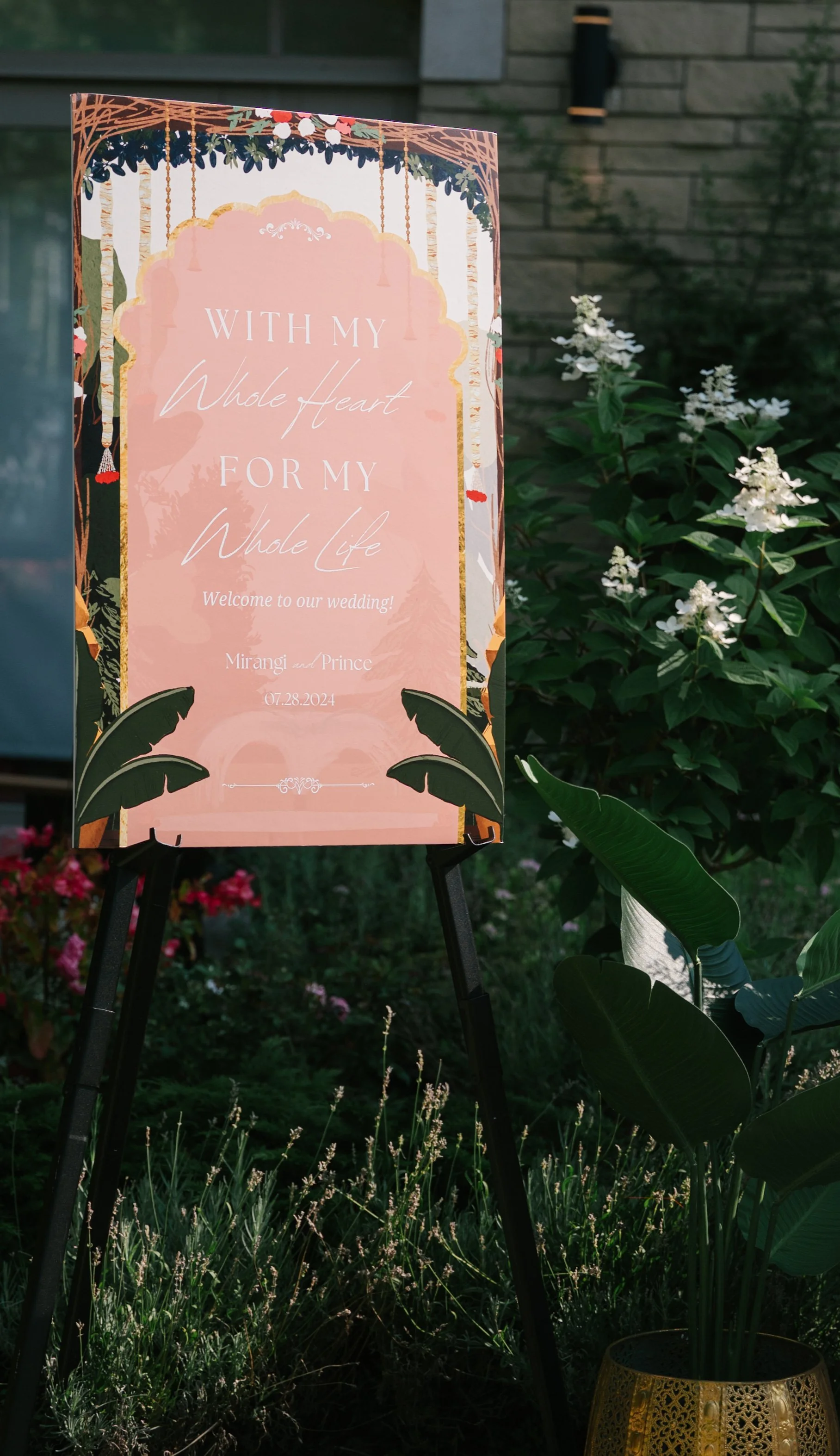 Wedding welcome sign with floral decorations, featuring the text 'With my Whole Heart for my Whole Life, Welcome to our wedding! Mirangi and Prince 07.28.2024,' surrounded by garden plants.