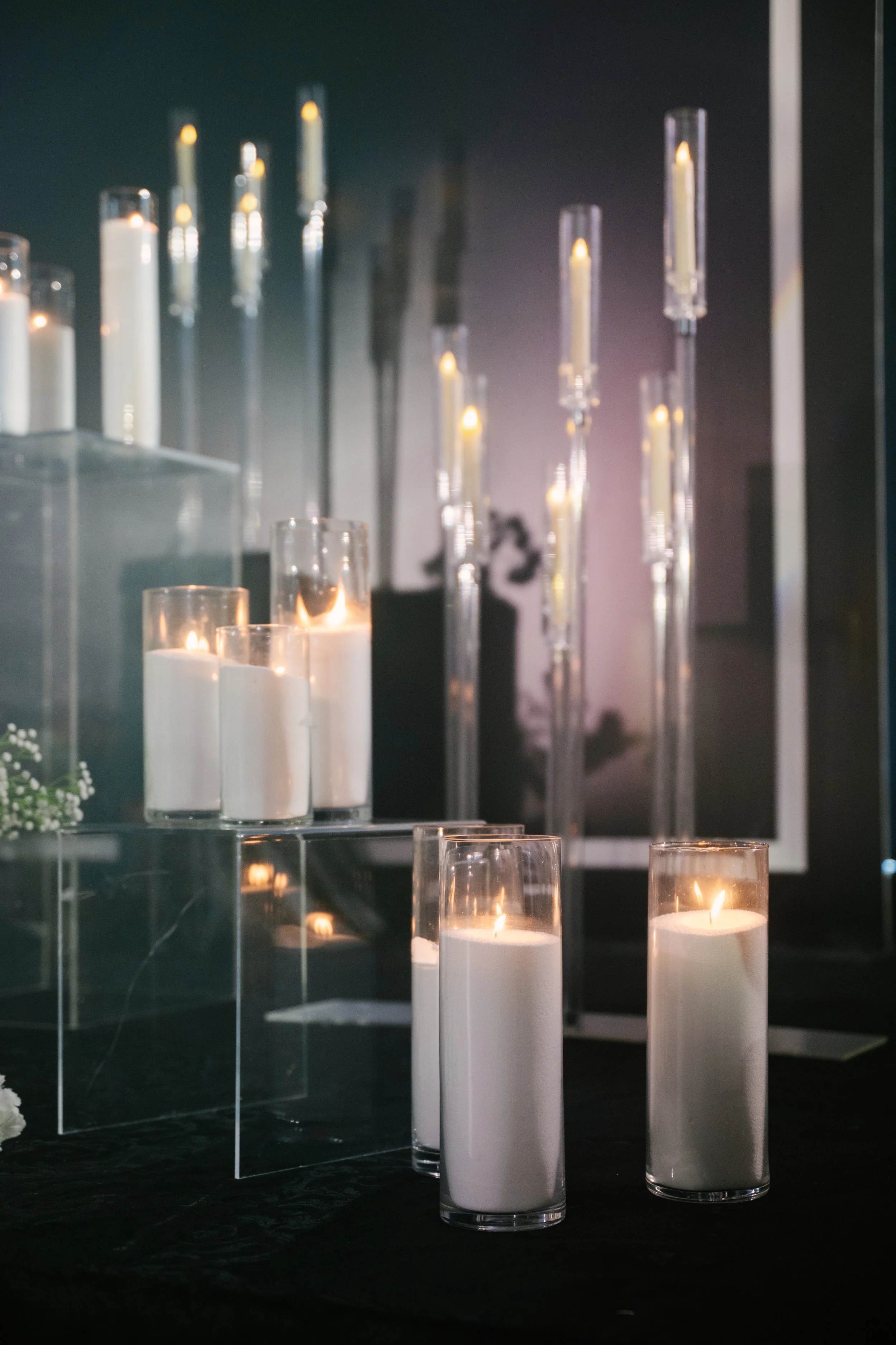 Lit white candles in glass holders arranged on reflective surfaces with a dark background.