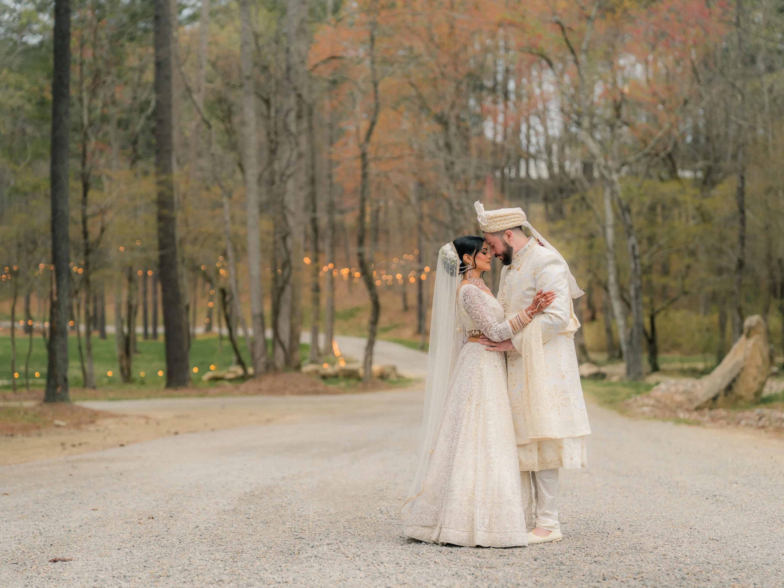 A couple dressed in traditional South Asian wedding attire stands closely on a tree-lined path. The bride is wearing an ornate white dress with a veil, and the groom is in a matching sherwani and turban. Ambient lights decorate the background among the trees.