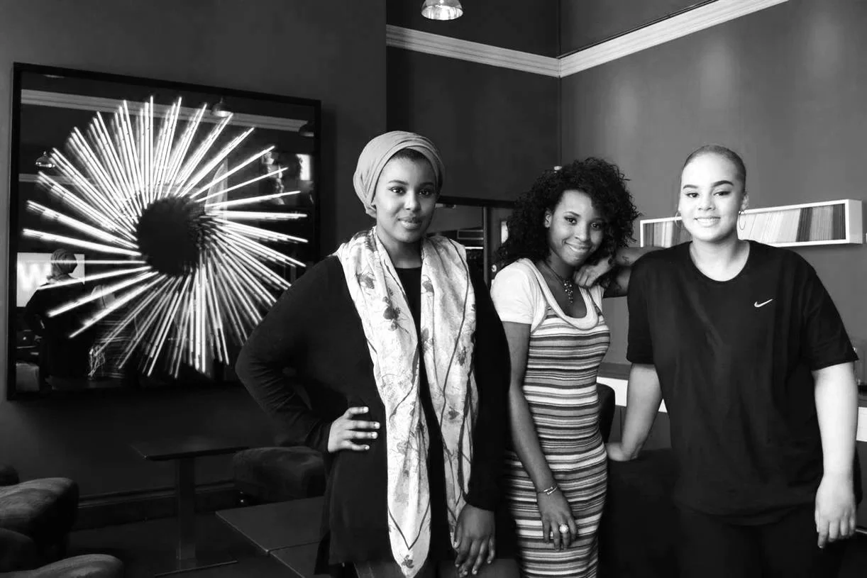 Black & white photo of 3 participants from The GirlHood Creative traineeship who were hired as junior creatives by the agency. They are smiling next to the agency logo.
