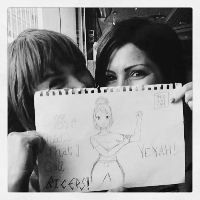 Black & white photo of Kati Russell and Natalie Rodden, co-founders of The GirlHood, peeking out from behind a drawing created by a workshop participant. The picture depicts a girl with big muscles and a slogan that reads Now that's what I call bicep