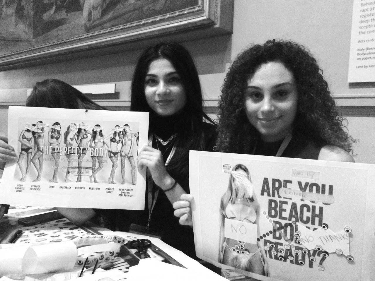 Black & white photo of two girls holding up two misogynistic ads which they have subverted using craft materials and gender positive language. Take during a workshop at the V&A.