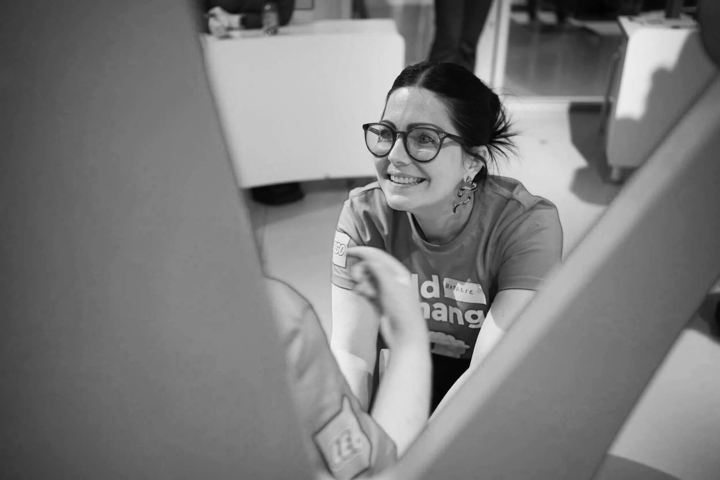 Black & white photo of Natalie talking to a student (out of view) and smiling at a LEGO Build the Change Youth Council event at the LEGO Discovery Centre in Birmingham.