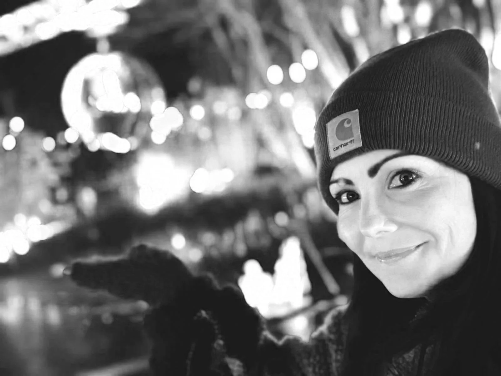 Black and white image of Natalie outdoors, gesturing to a disco ball in the distance. She's smiling in warm winter clothing and a beanie hat.