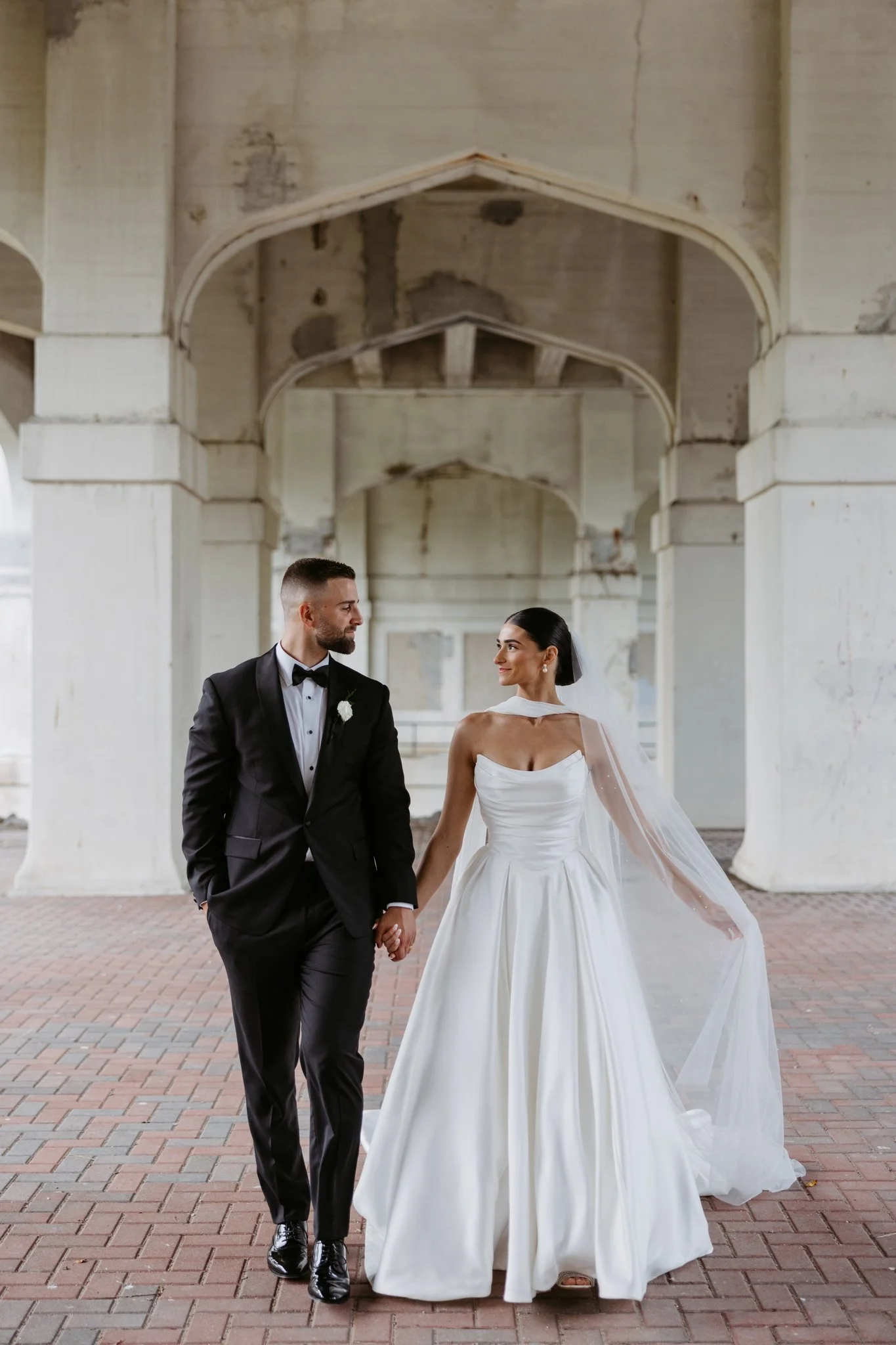 Hannah and Atticus holding hands beneath a stone archway during their Petoskey wedding portraits. Photography by Amelie Ferdais.