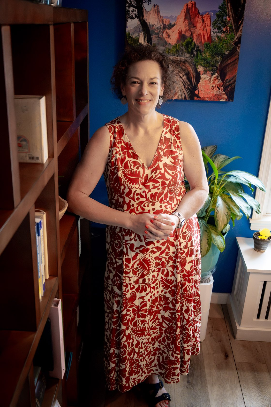 A woman with curly hair and earrings, wearing a sleeveless red and white floral dress, standing indoors near a bookshelf and a potted plant, with a landscape painting of red rocks and trees on a blue wall behind her.
