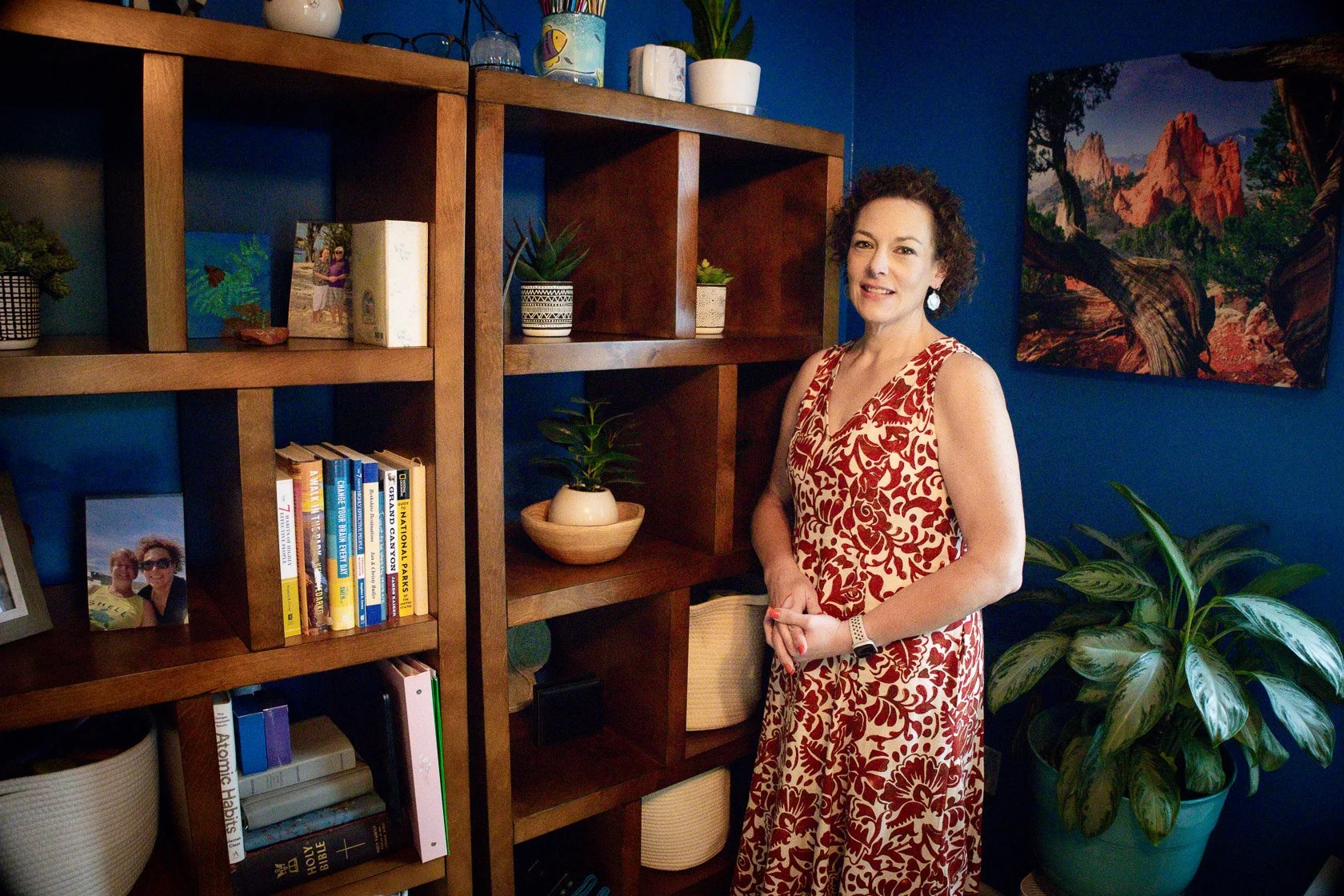 A woman with curly hair wearing a red and white patterned dress standing next to a bookshelf with books, framed photos, and plants against a blue wall in a living room.