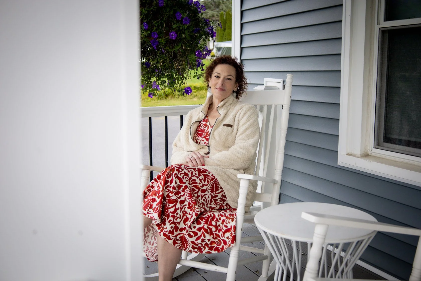 A woman sitting on a white rocking chair on a porch, with purple flowers hanging in the background and blue siding on the house.