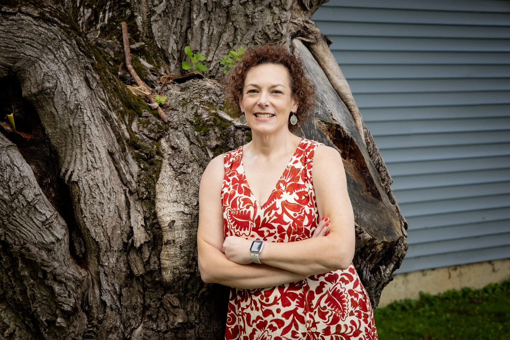 A woman with short curly hair, wearing a red and white floral dress, standing outdoors with her arms crossed, smiling in front of a large tree trunk and a gray building with horizontal siding.
