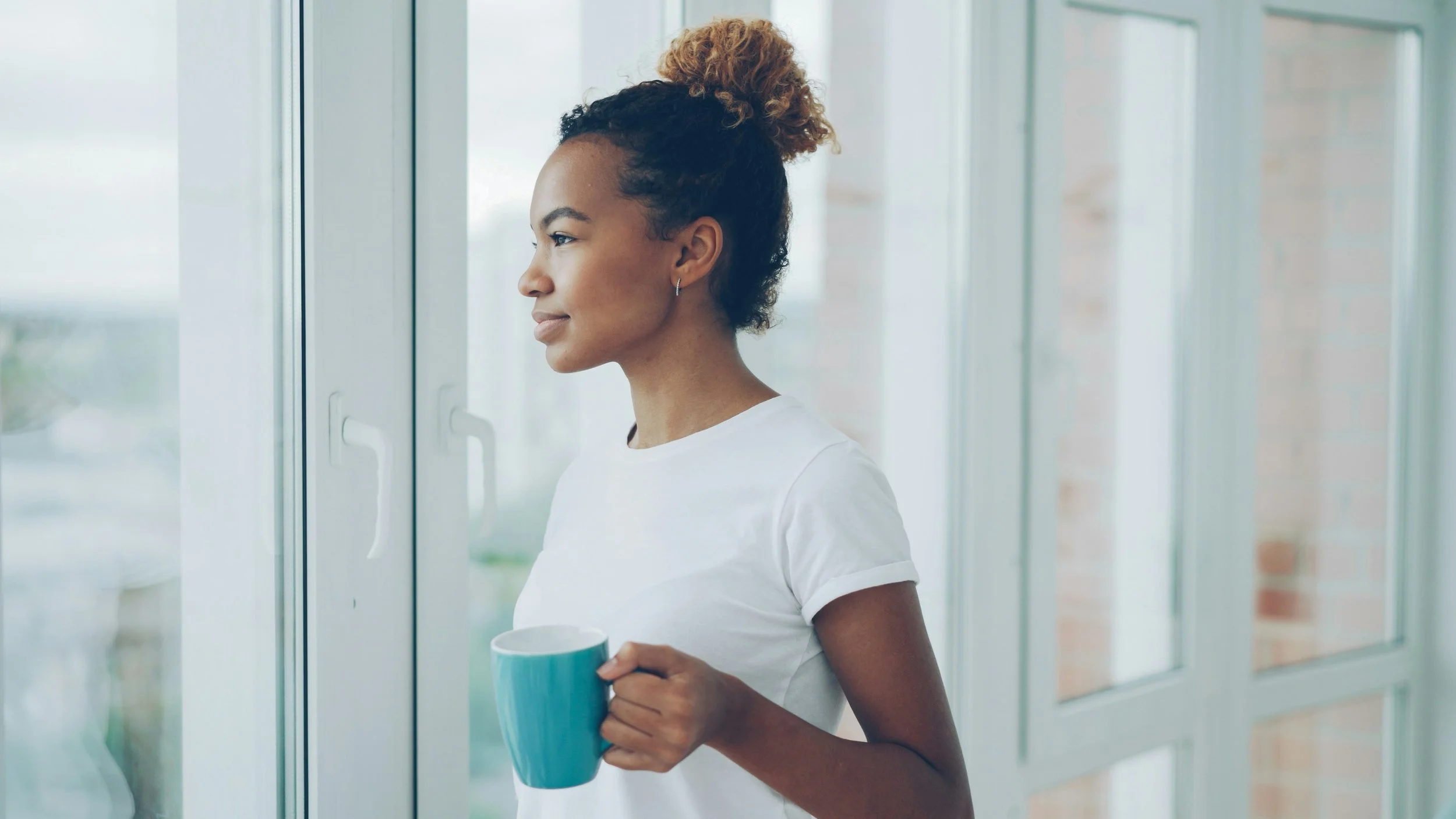 A woman with curly hair in a high bun, wearing a white shirt, looking out a window while holding a blue mug