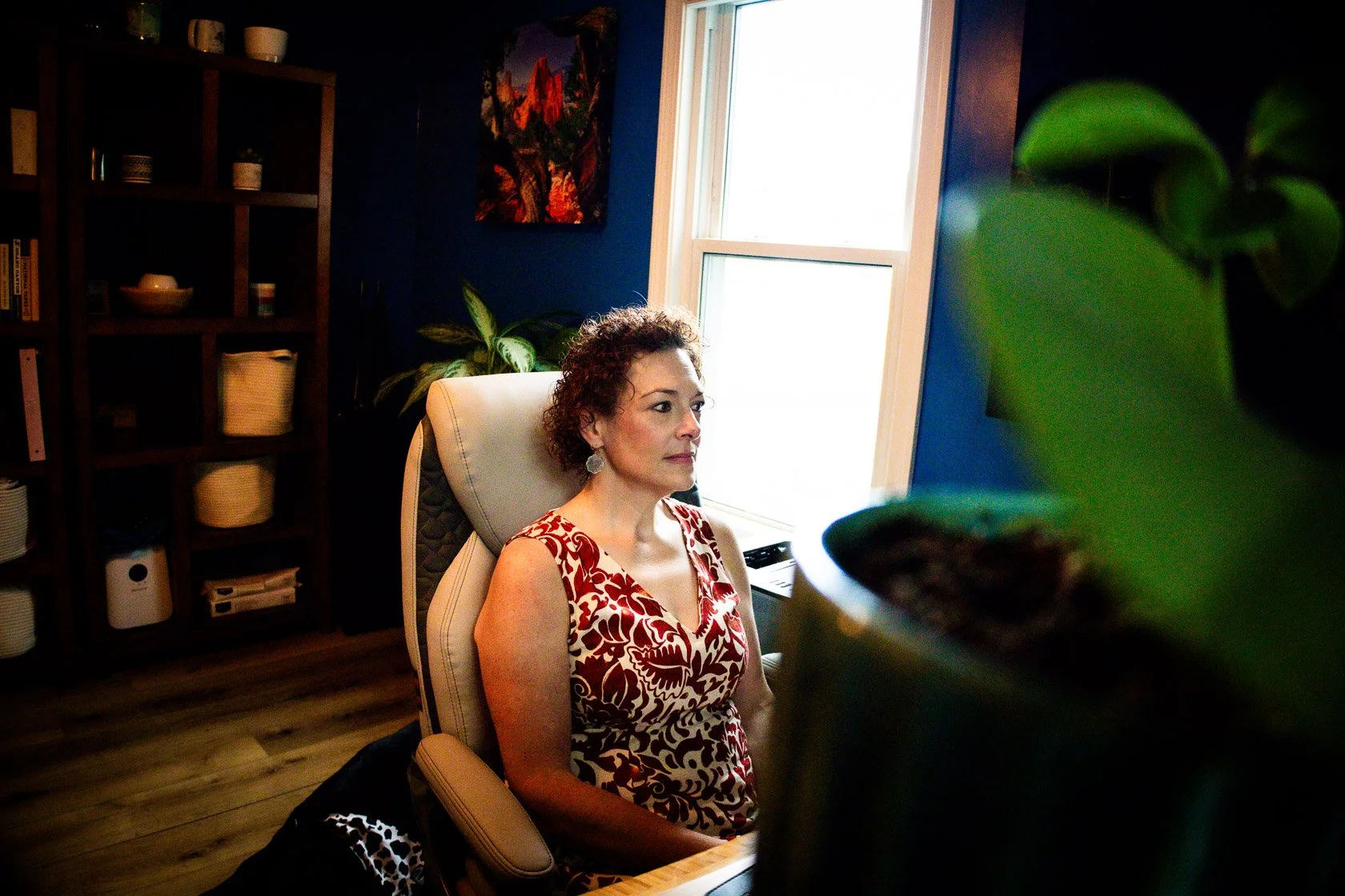 A woman with curly hair sitting in a beige office chair in a room with dark blue walls, near a large window, with a bookshelf and a vibrant painting in the background.