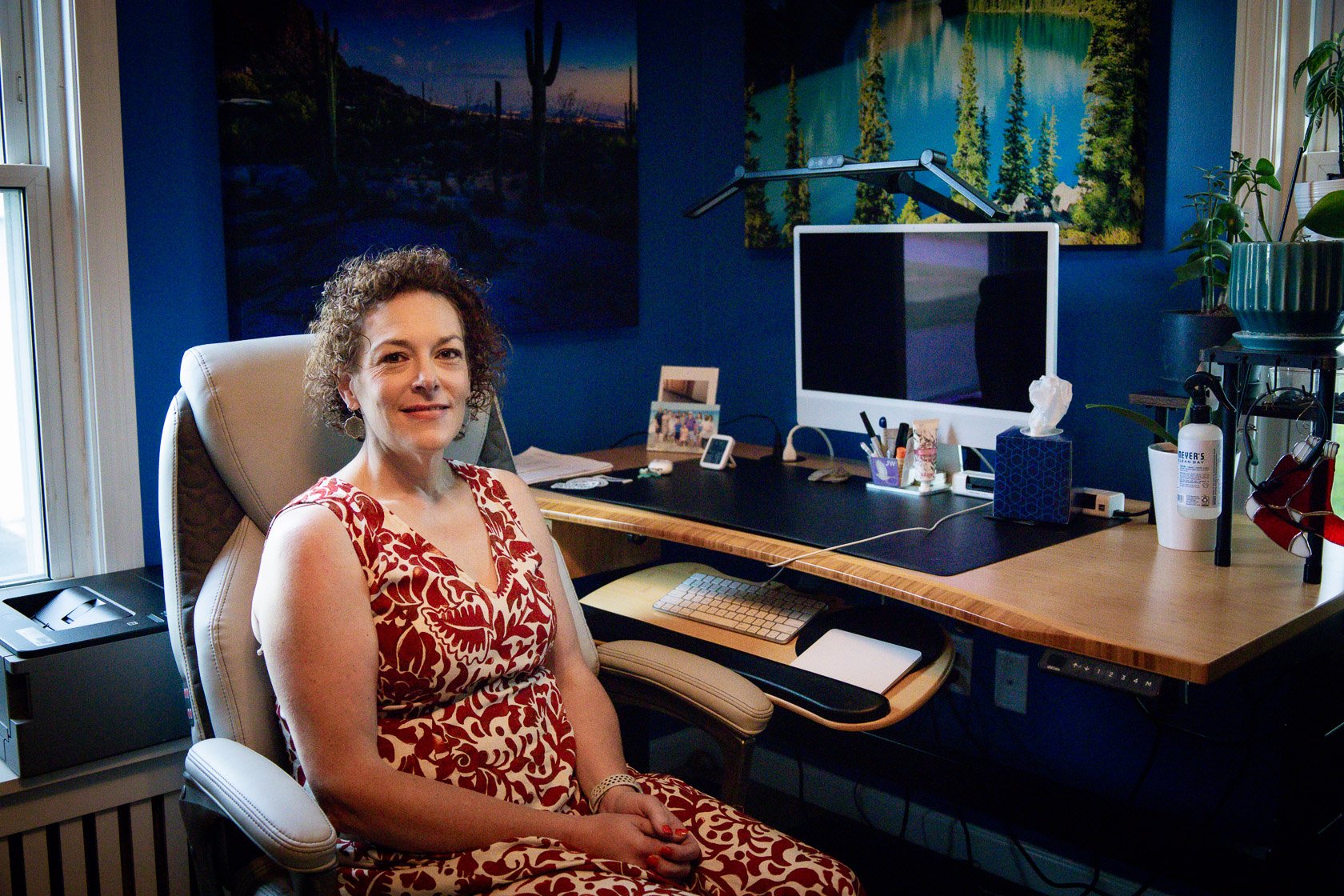 A woman in a red and white patterned dress sitting in an office chair at a desk with computer equipment, plants, and artwork on blue walls.
