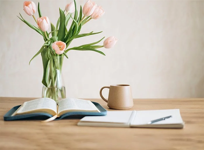 A vase with pink tulips, an open book, a ceramic mug, and a notebook with a pen on a wooden table.