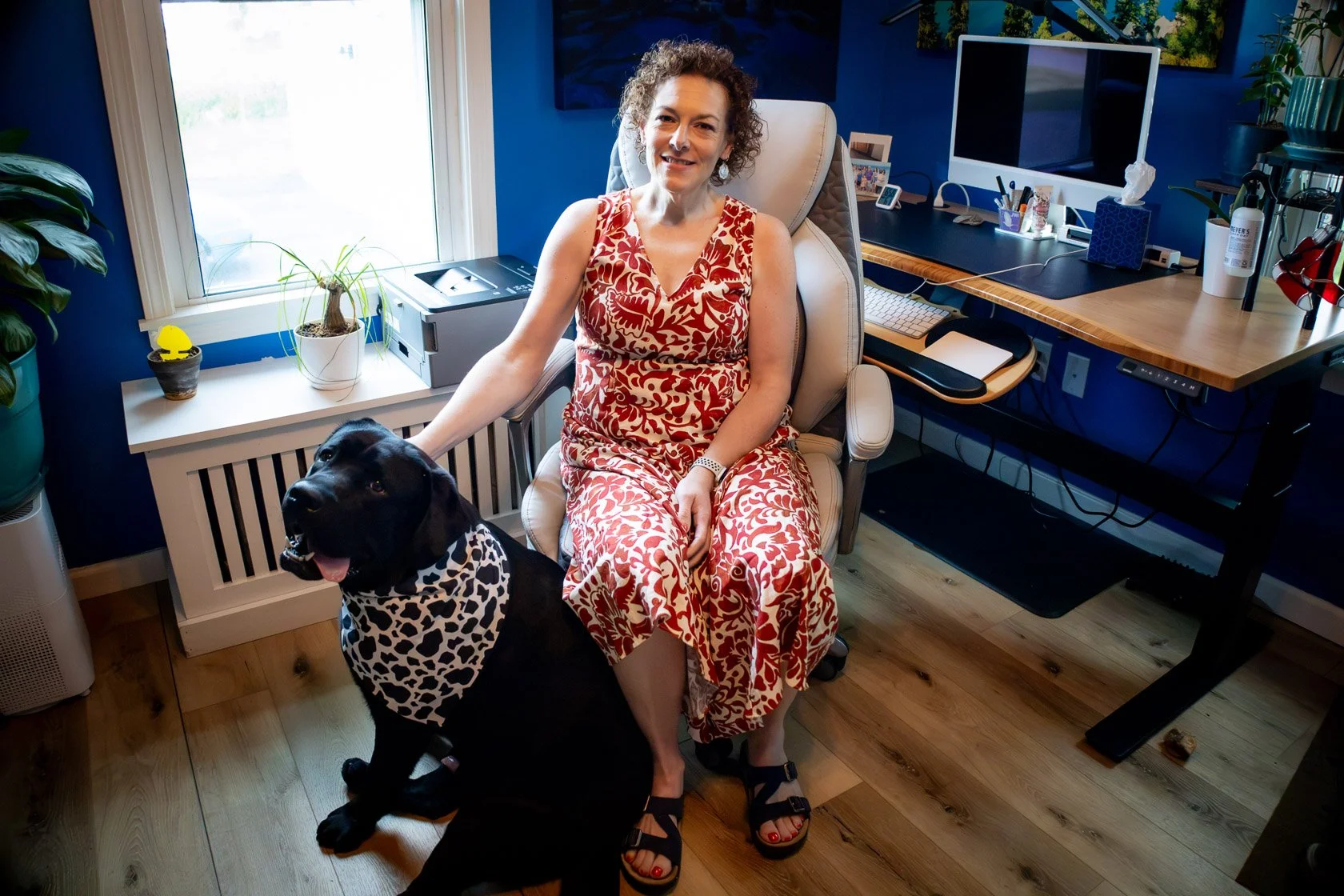 A woman with curly hair in a red and white patterned dress sitting on an office chair, petting a black dog with a leopard print bandana, in a home office with a desk, computer, and plants.