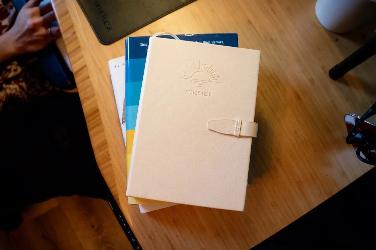A beige planner with a strap closure and the words 'Stress Less' embossed on the cover, resting on a stack of other books on a wooden desk.