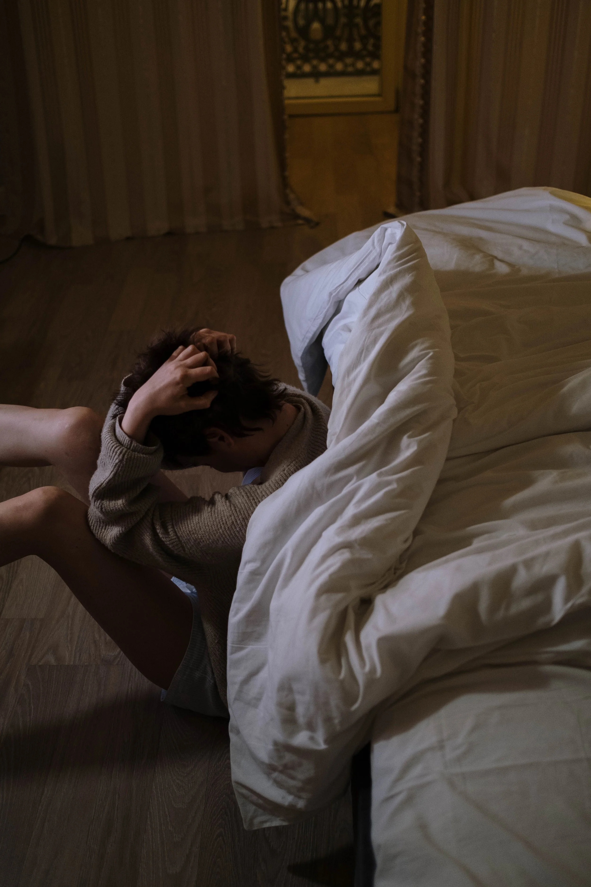 A person with short hair and light skin laying on a bed with white sheets, holding their head with both hands, in a dimly lit room with striped curtains and wooden floors.