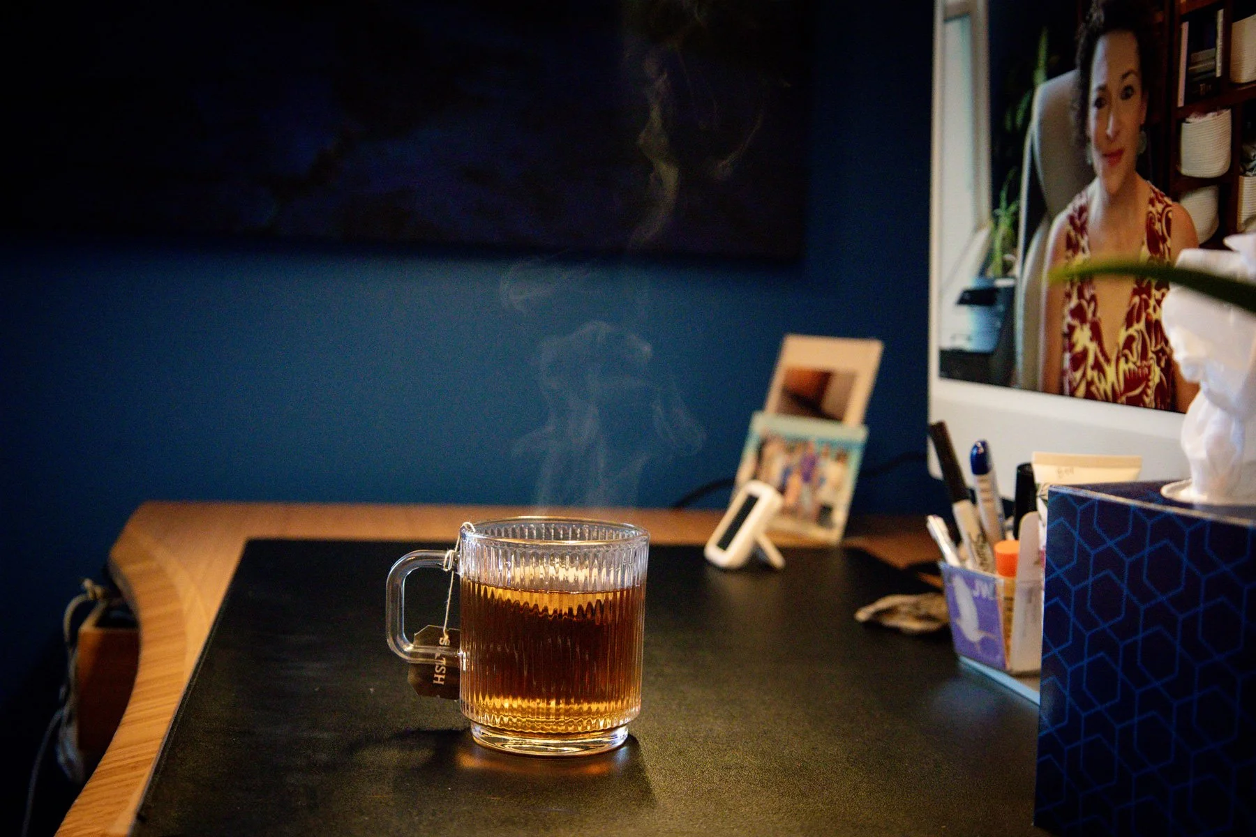 A glass mug filled with hot tea sitting on a dark surface, with a wooden edge visible. In the background, a mirror reflects a woman with curly hair and a red and yellow patterned dress, sitting in a well-lit room with shelves and plants. There are various objects on the table, including a tissue box, a framed picture, pens, and other small items.