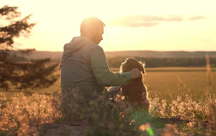Person sitting outdoors with dog during sunset in a natural landscape.