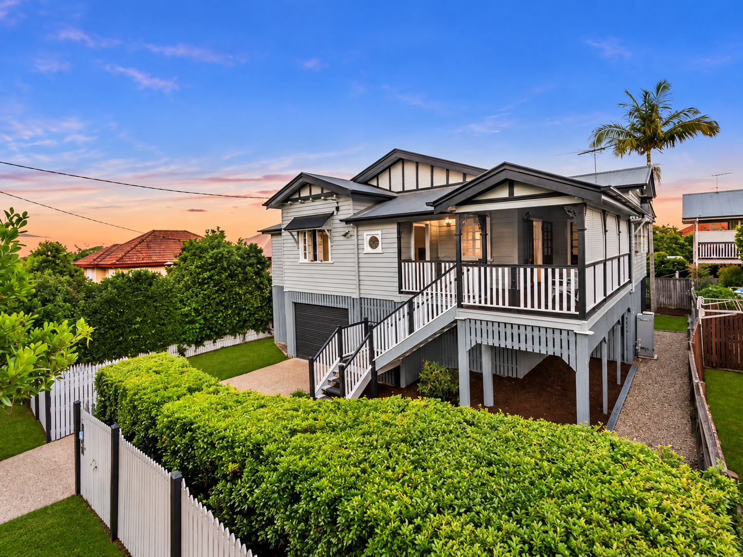 Raised Queenslander home with a front verandah, timber stairs and landscaped garden at dusk.