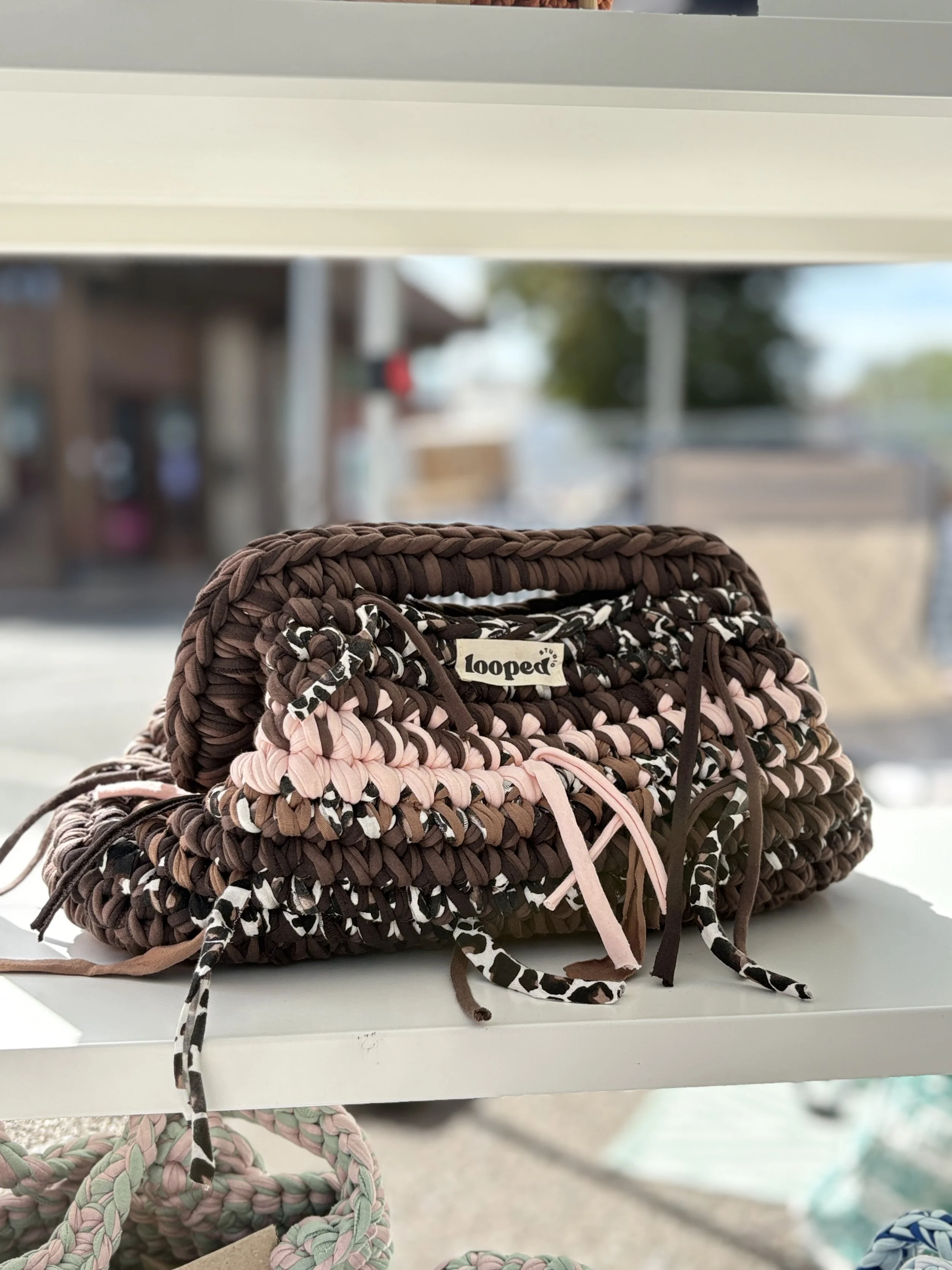 Close-up of a woven bag with a 'looped' brand tag, placed on a white surface with other similar woven bags in pastel colors around it. The background shows a blurred outdoor scene with structures and trees.
