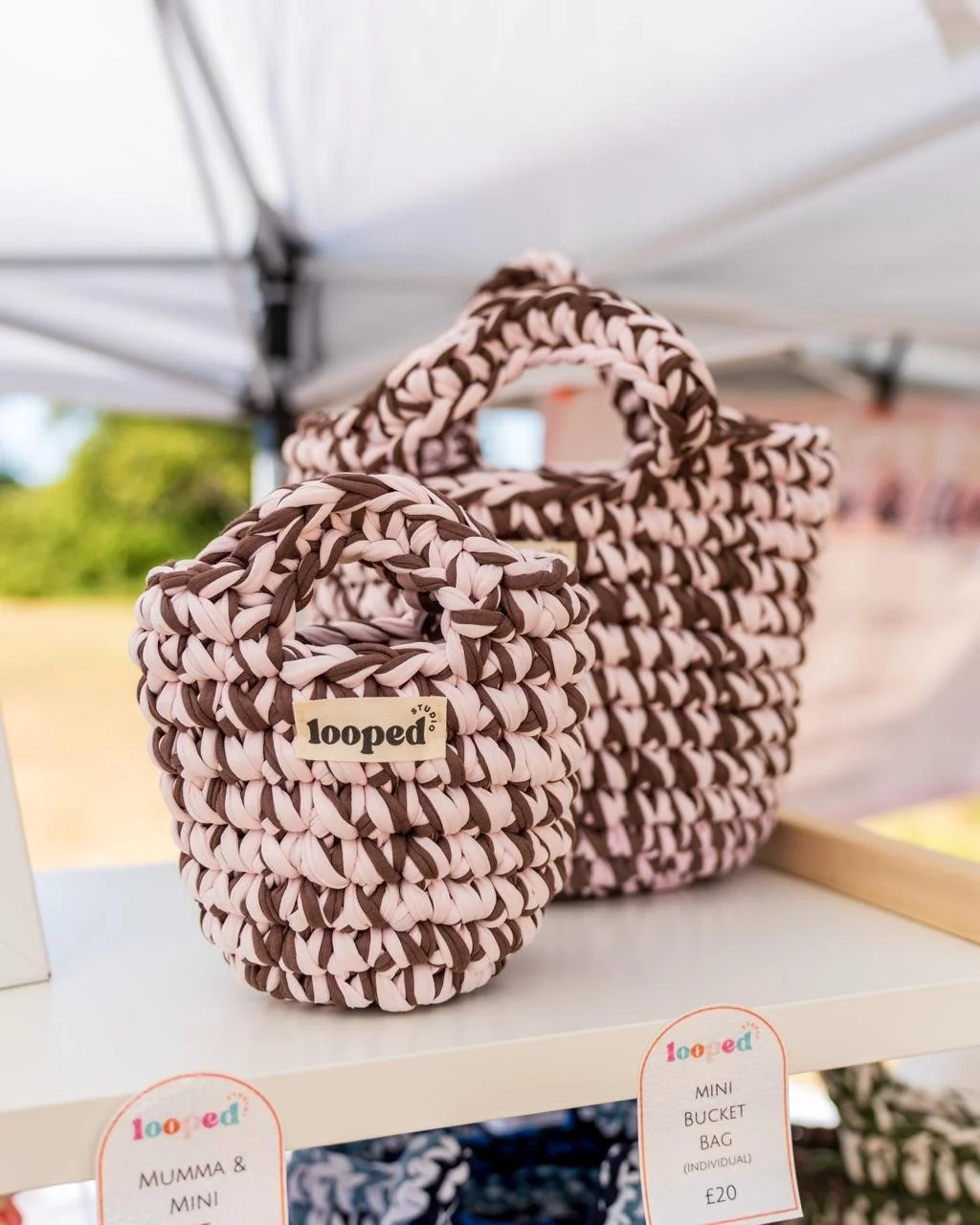 Two handwoven mini bucket bags made from pink and brown fabric, displayed on a white shelf under a tent at an outdoor market.