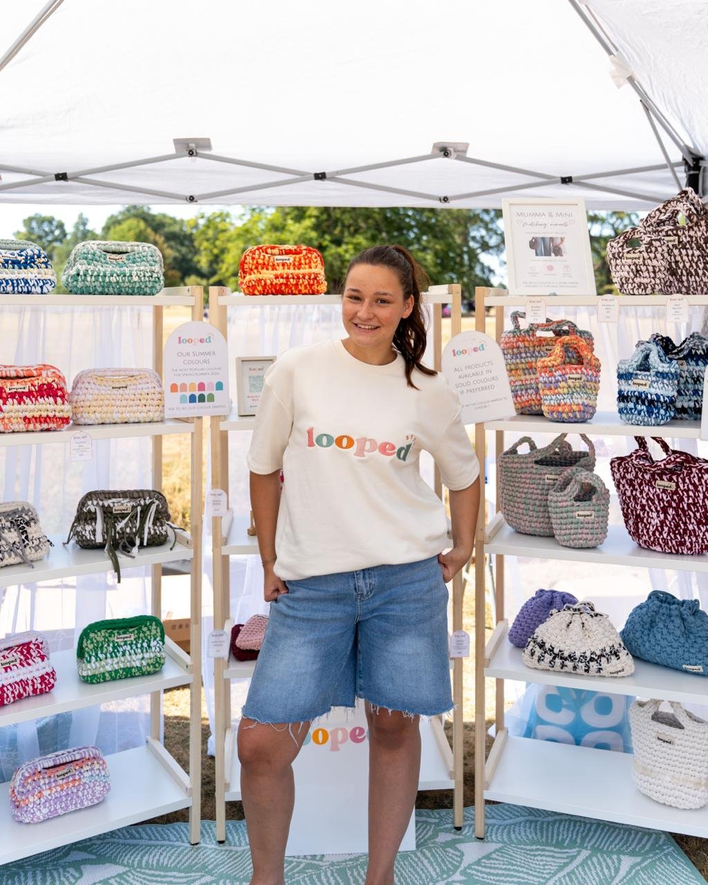 Smiling woman standing in front of a display of colorful crocheted bags at an outdoor market.