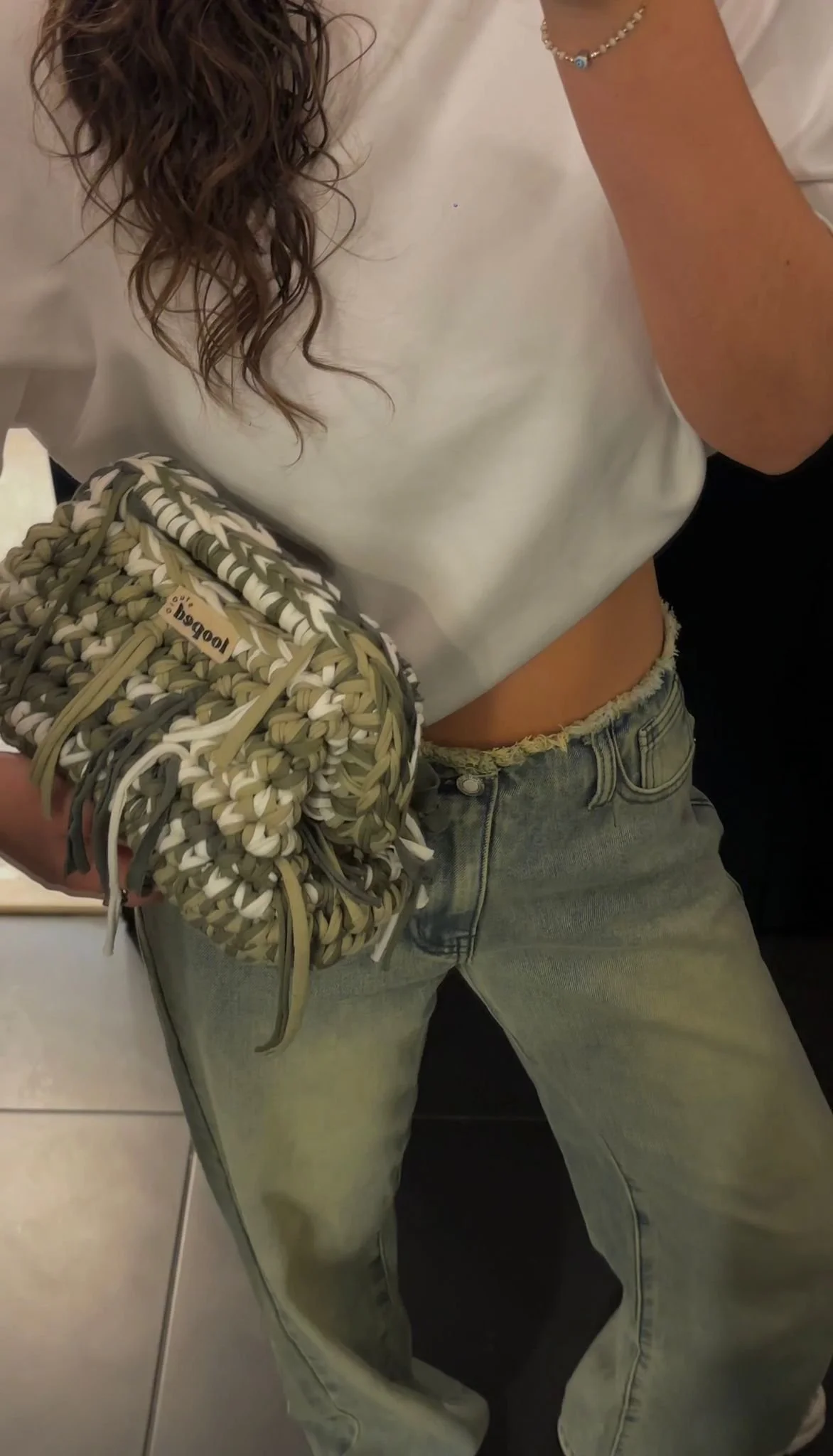 Person wearing a white top, light blue jeans with frayed waistband, holding a large woven bag with tassels, standing in an indoor setting.