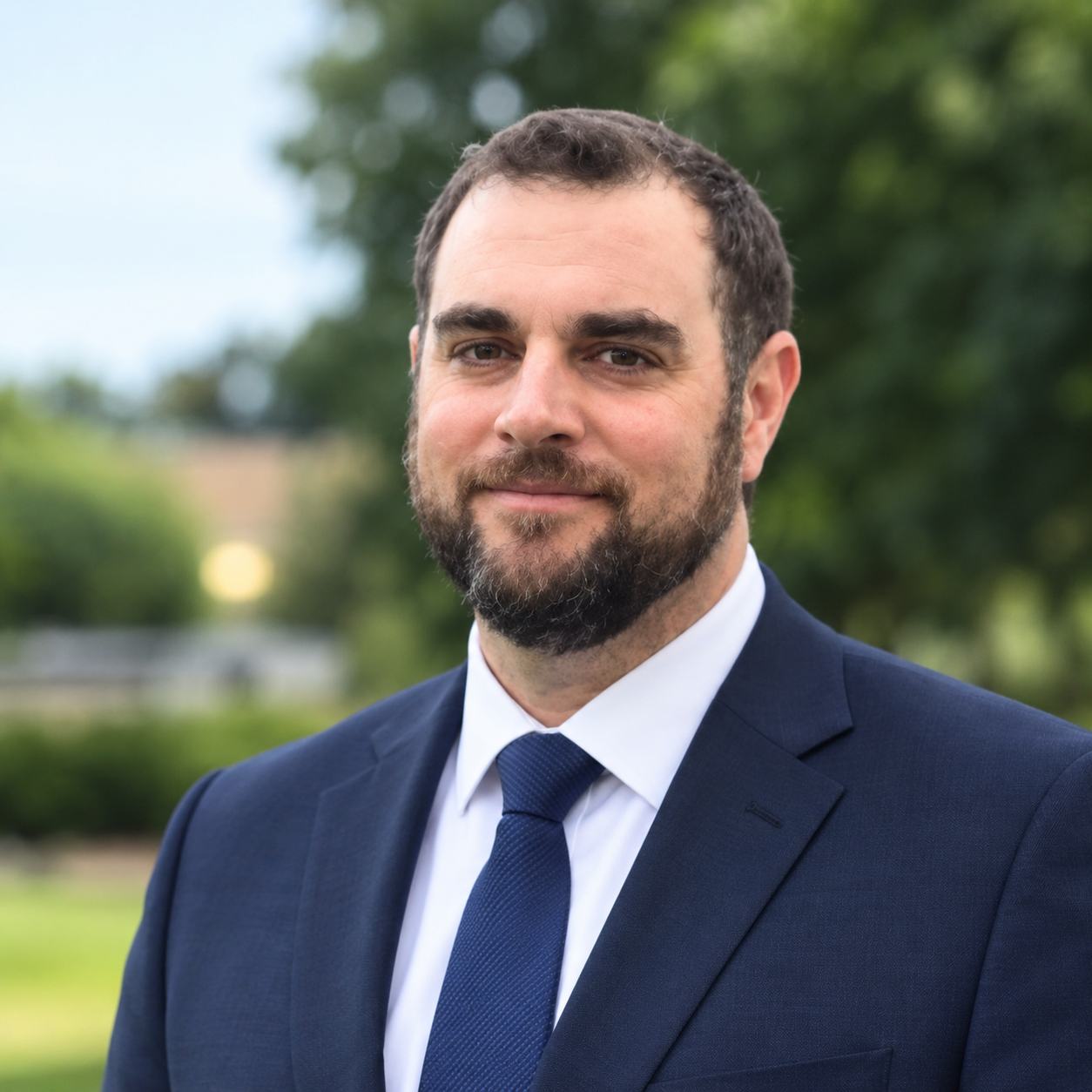 A man with a beard in a navy suit and tie standing outdoors with blurred trees and foliage in the background.