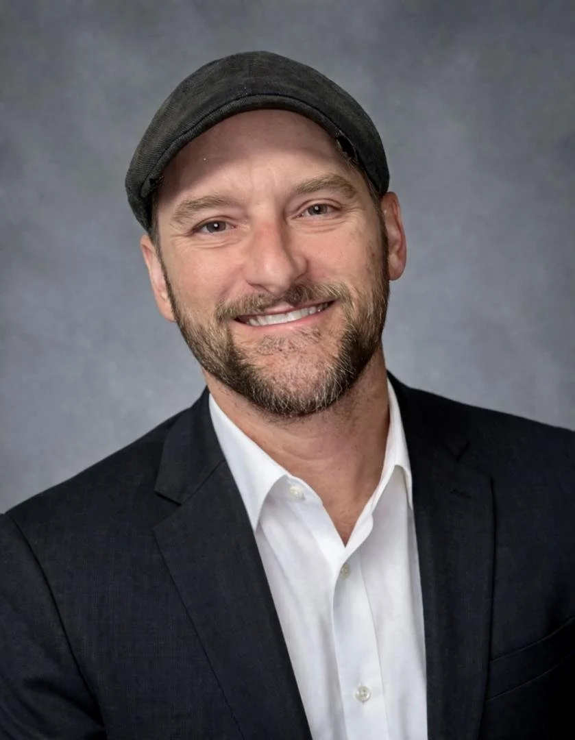 A professional man with a beard wearing a black cap, white shirt, and black blazer, smiling at the camera, against a gray background.