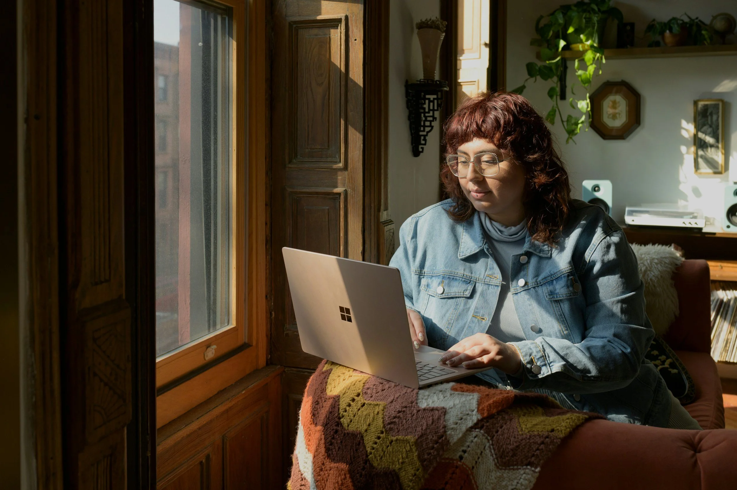 Woman with curly hair and glasses working on a laptop near a wooden window with sunlight streaming in.