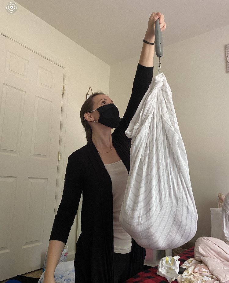 A woman wearing a black face mask and black cardigan is reaching up to lift a large white cloth bag with a handle, in a room with light-colored walls and a closed door.