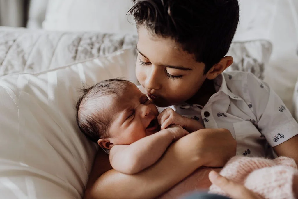 A young boy gently holds a newborn baby in his arms, both lying on a bed, with the boy's face close to the baby's.