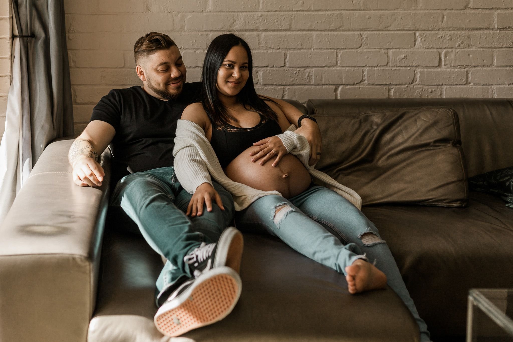 A couple sitting on a brown leather couch in front of a brick wall. The woman, who is pregnant, is sitting between the man's legs with her hands on her belly. The man has his arm around her, and they are both smiling, looking cozy and happy.