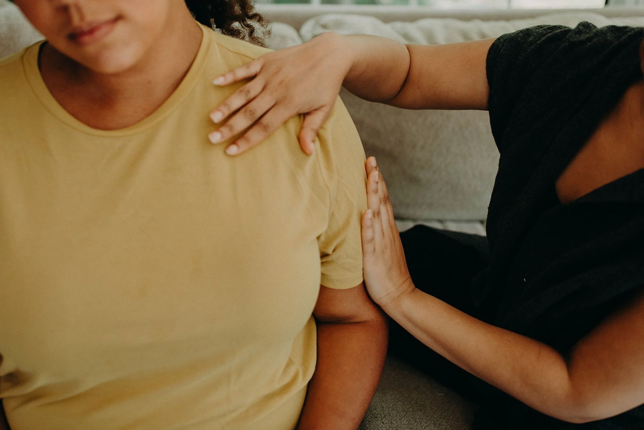 A person wearing a black shirt is placing their hand on the shoulder of another person wearing a yellow shirt. The second person's face is partially visible and they are smiling. The scene appears to be indoors with a sofa or bed in the background.