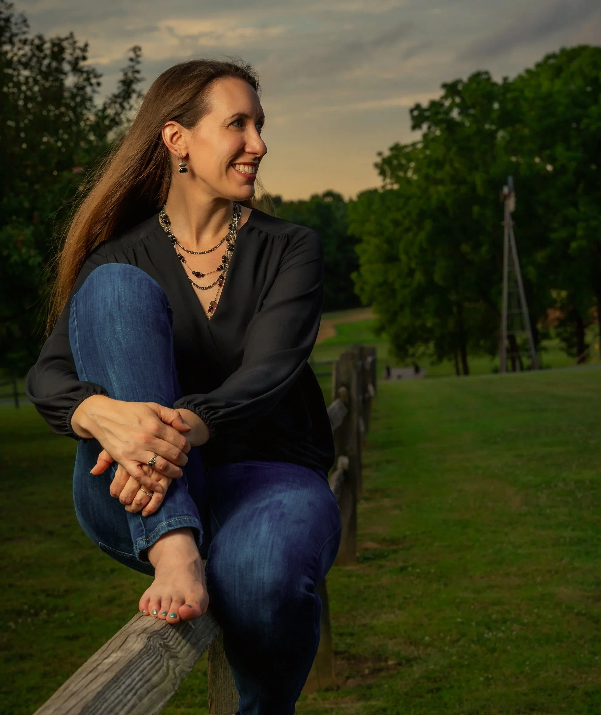 A woman with long brown hair, wearing a black top and blue jeans, sitting on a wooden fence outdoors during sunset. She is smiling and looking to her left, with trees and a cloudy sky in the background.