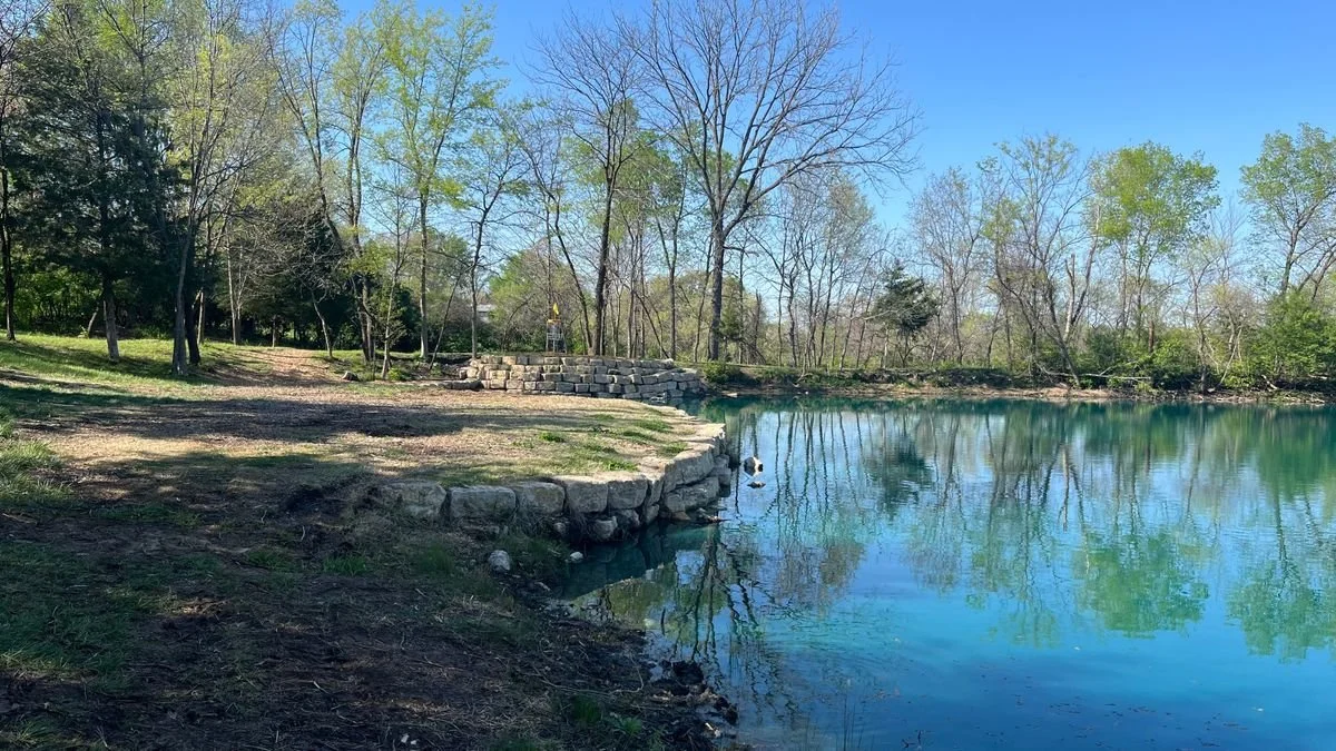 Peaceful lakeside scene with a stone embankment, leafless and green trees, reflecting in calm water, under a clear blue sky.