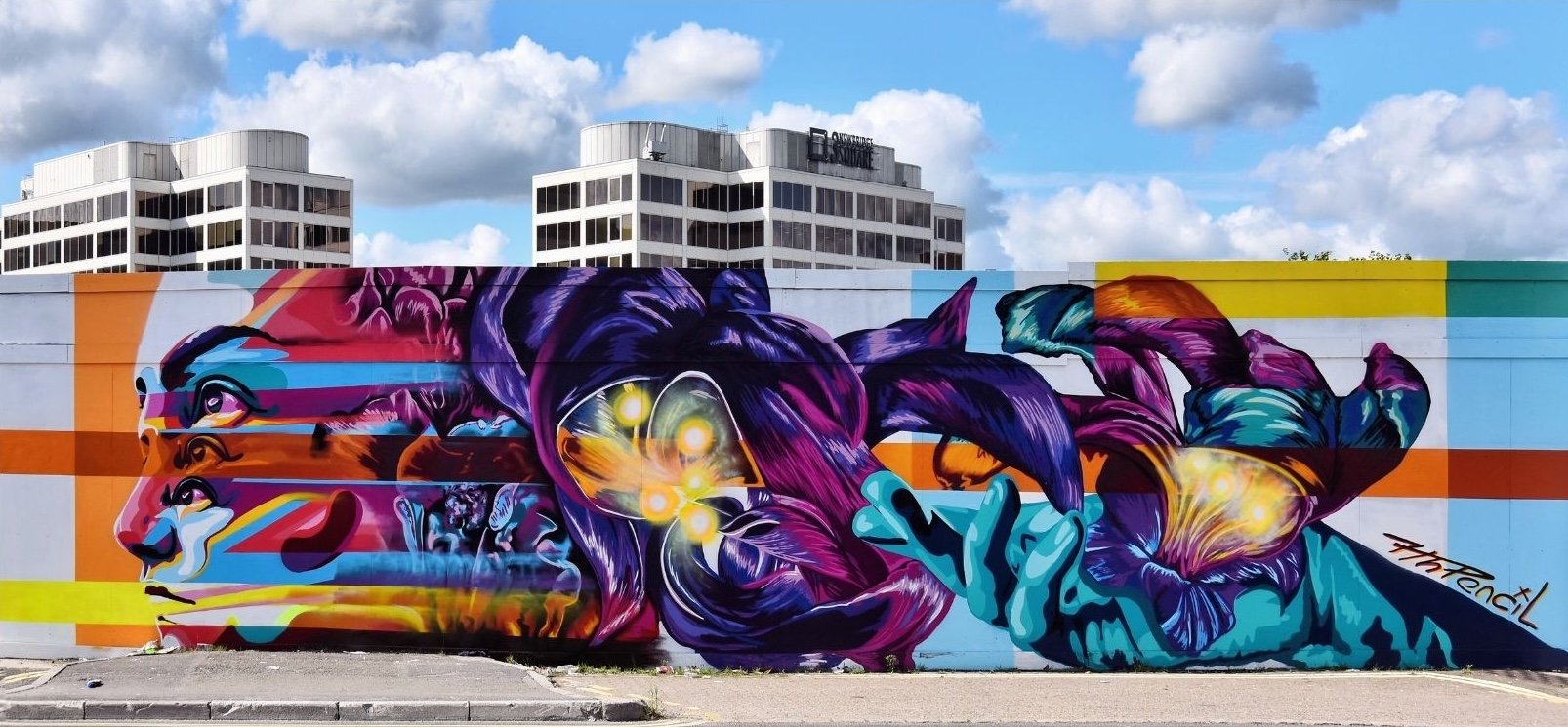 Colorful graffiti mural on a wall featuring abstract human faces, purple leaves, and glowing orbs with blue and orange stripes, with city buildings and a partly cloudy sky in the background.